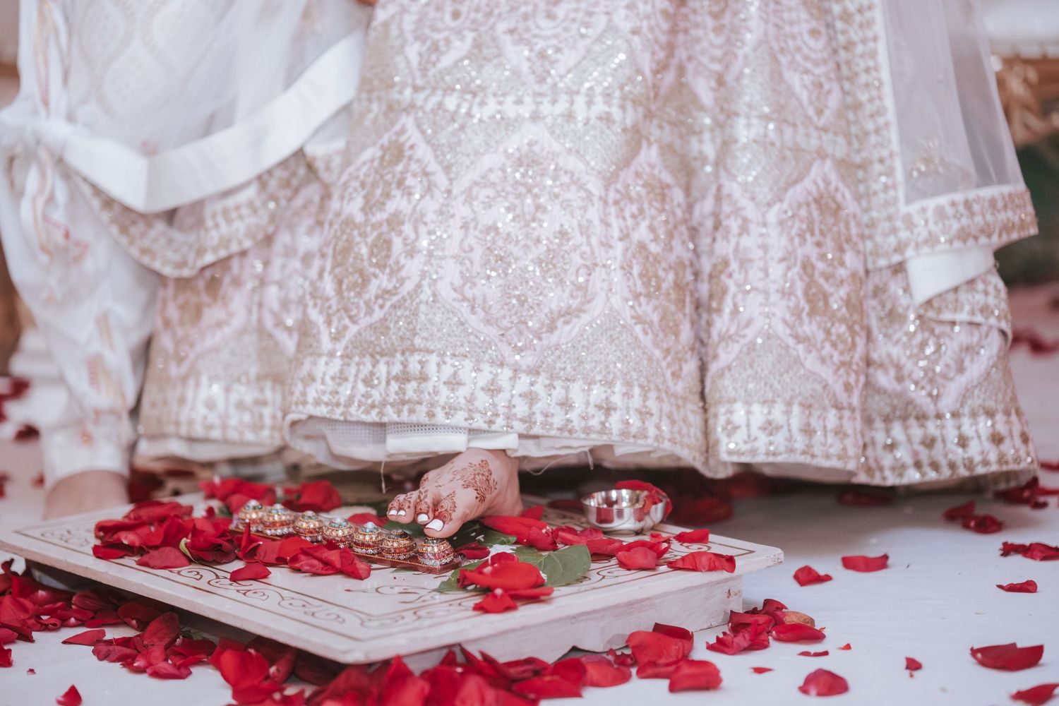 Close-up of Indian bride's feet near wedding ritual items surrounded by red rose petals on white floor.