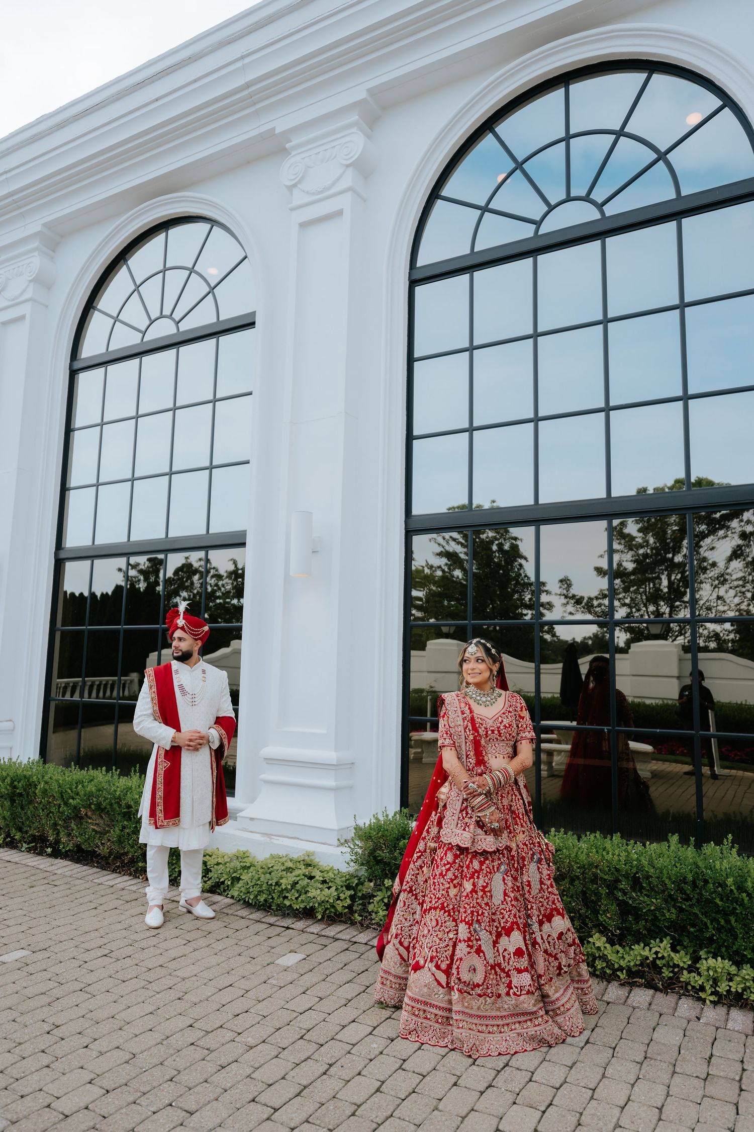 Indian wedding couple in red lehenga and white sherwani stand apart before grand arched windows.