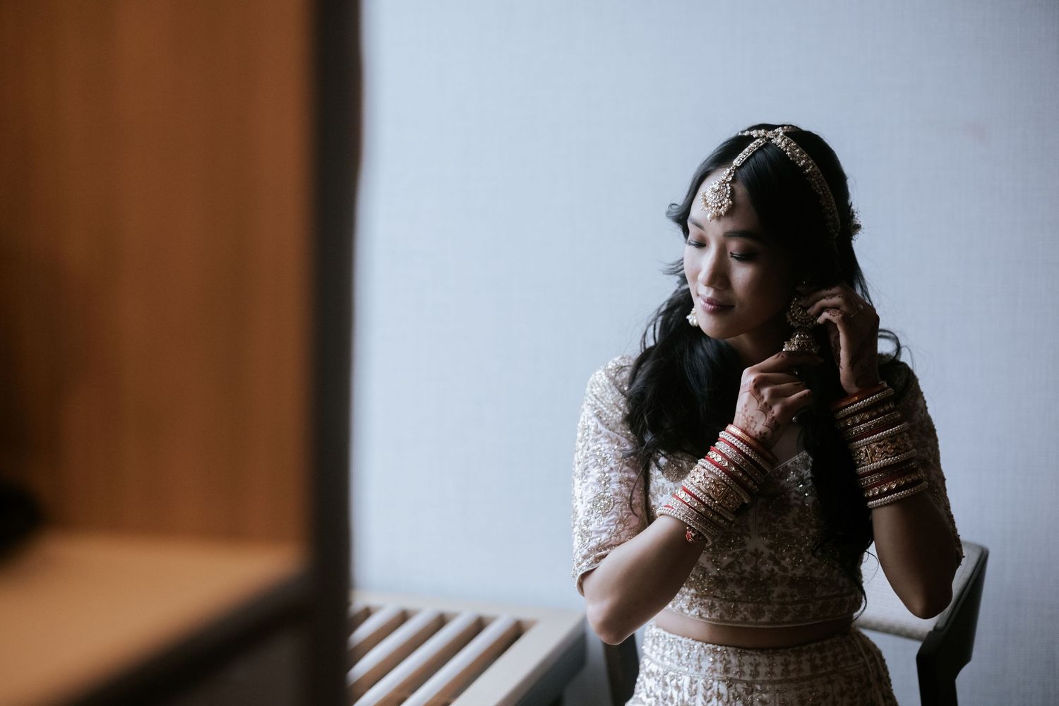 Indian bride in white and gold lehenga adjusting her earring while gazing out a window on her wedding day.