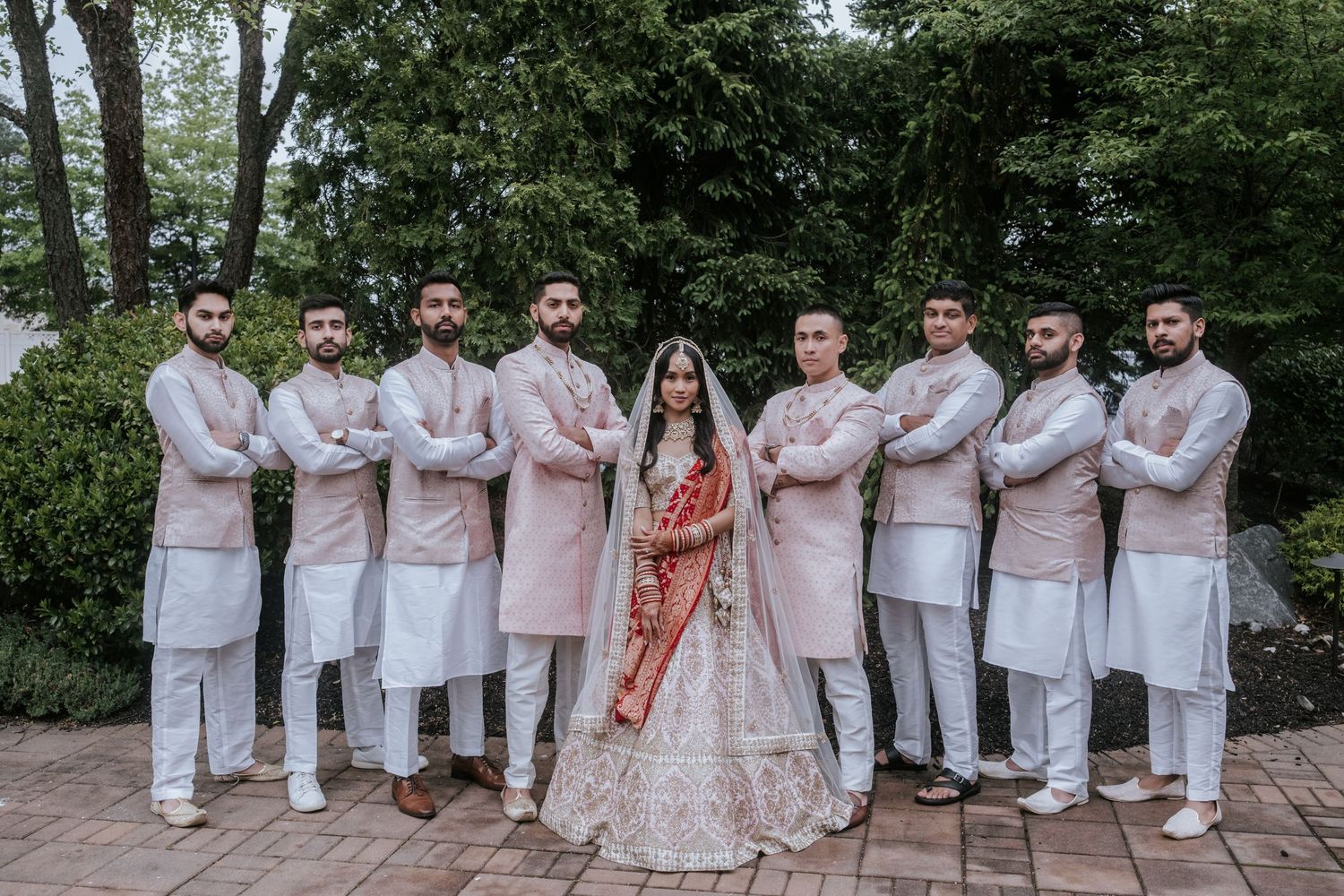 Indian bride in red lehenga surrounded by groomsmen in pink sherwanis posing with arms crossed outdoors.