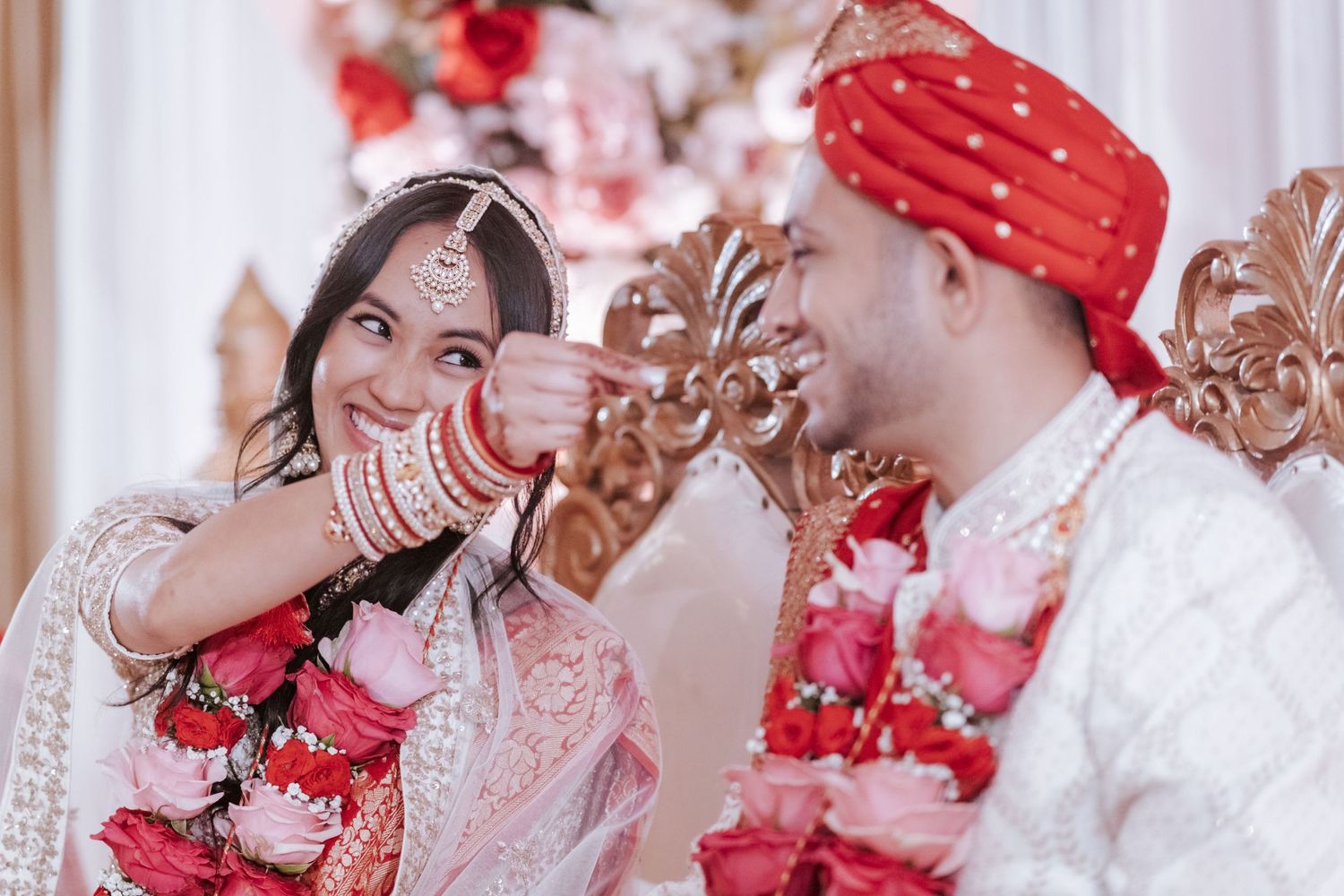 Indian bride and groom laughing joyfully during wedding ceremony, adorned with flower garlands and traditional attire.