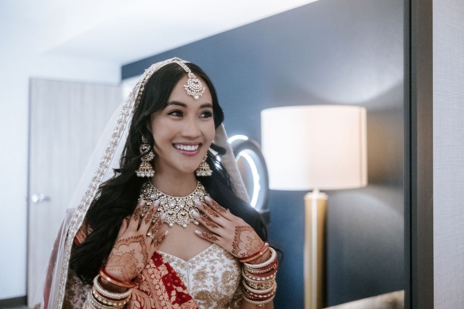 Radiant Indian bride in red and white lehenga with diamond necklace and dupatta smiles.