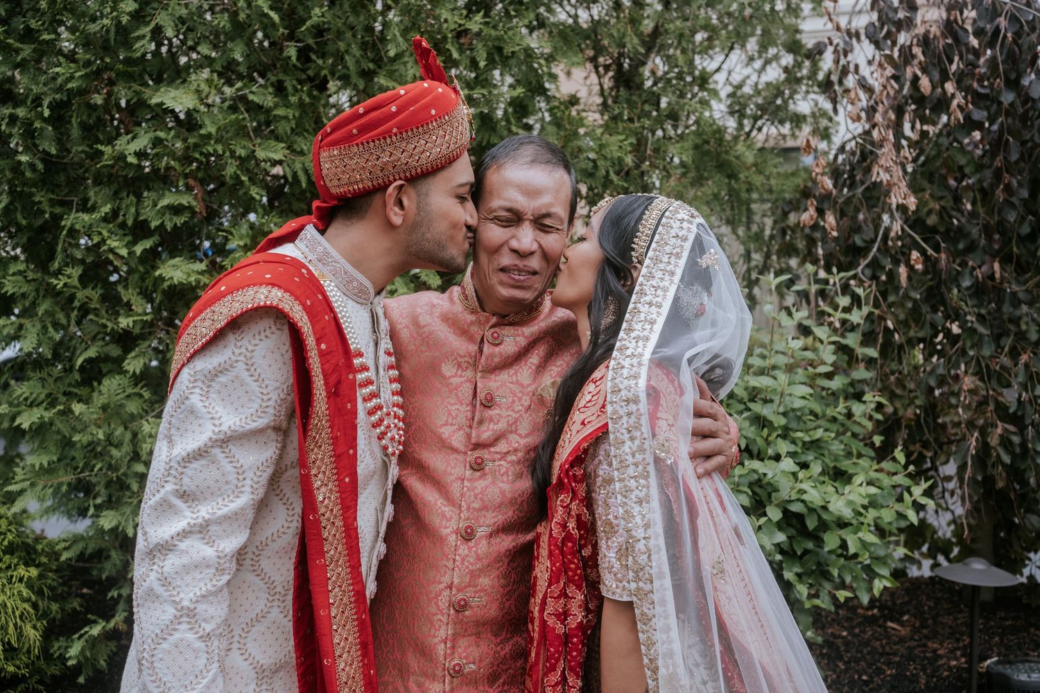 Indian bride and groom in traditional wedding attire kiss elderly man on cheeks in garden setting.