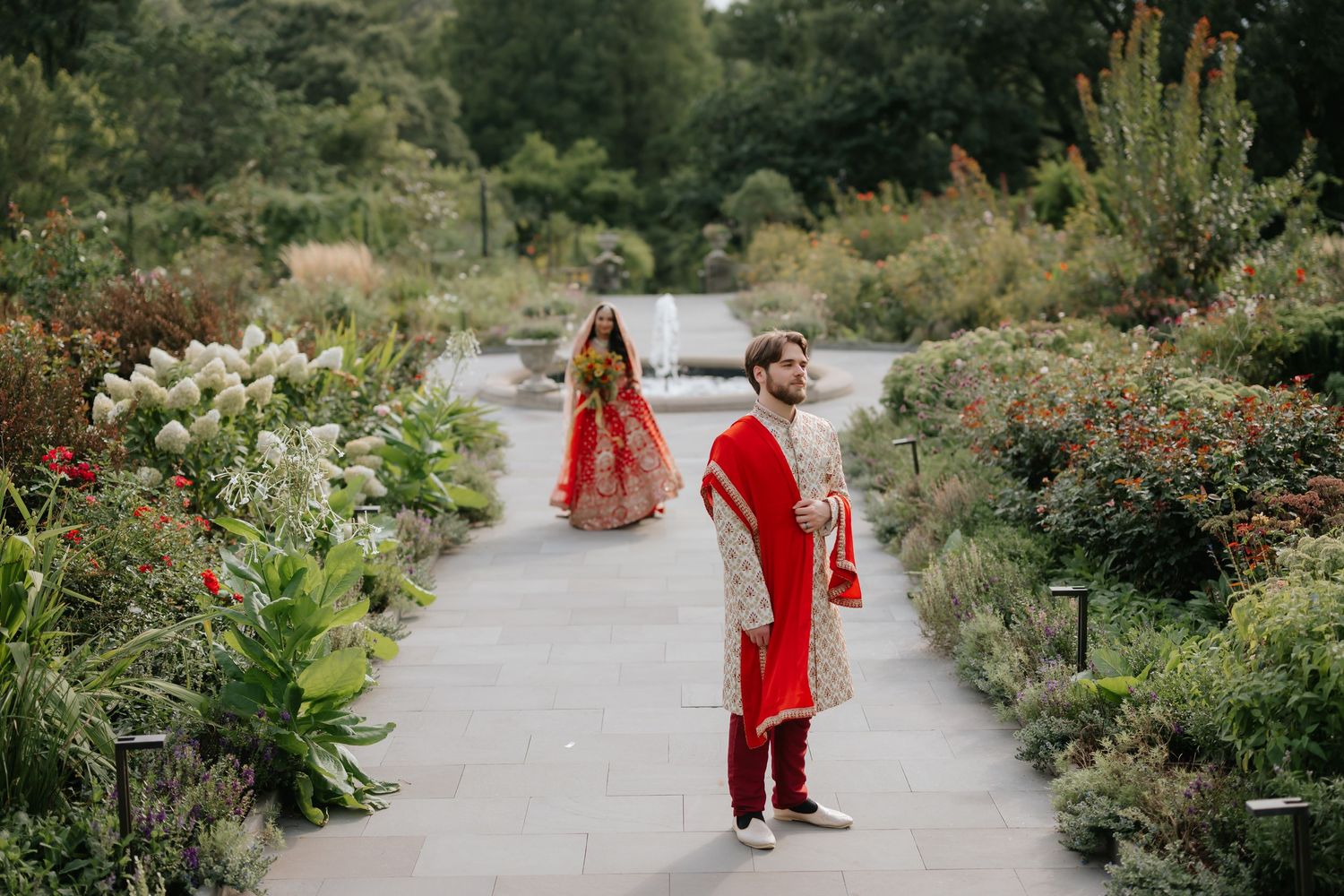Indian bride and groom in red and gold traditional attire standing back to back in garden path.