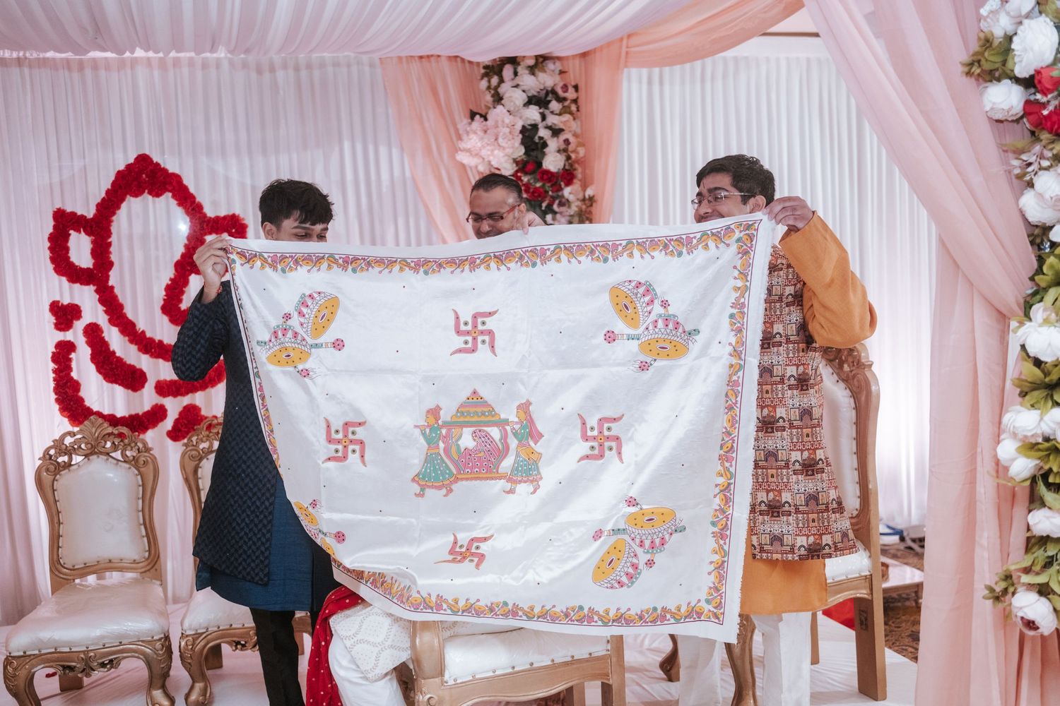 Two men holding embroidered ceremonial cloth with Hindu symbols at Indian wedding mandap.