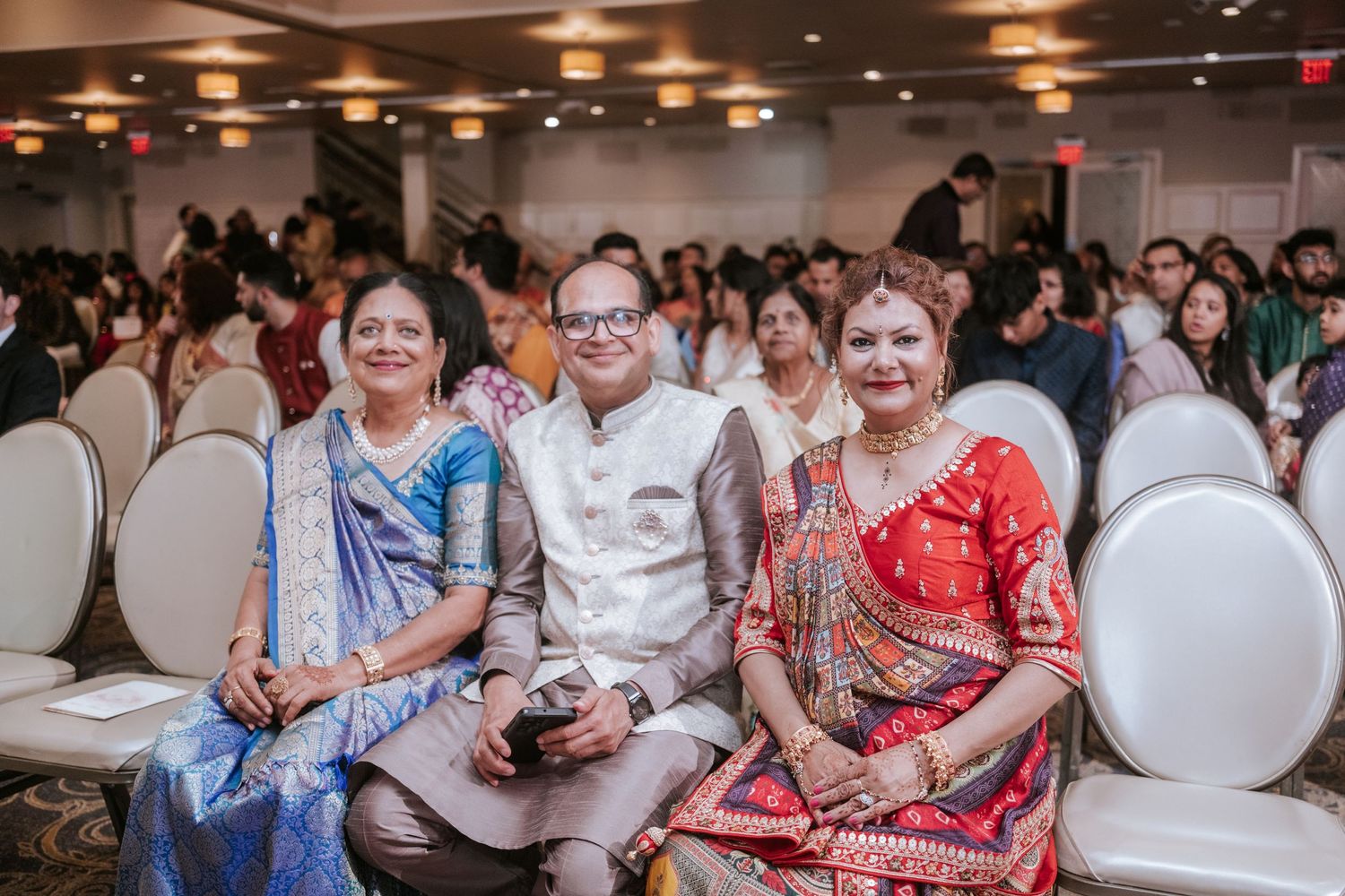 Three elegantly dressed guests in colorful sarees and kurta smile while seated at Indian wedding.