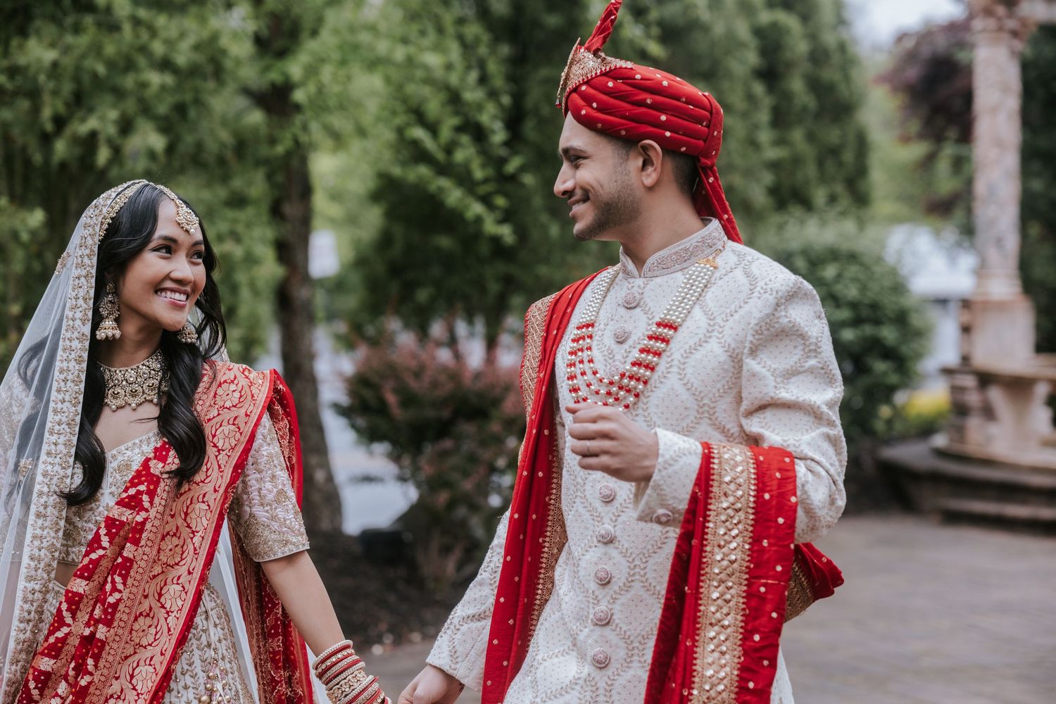 Indian bride and groom smiling at each other outdoors, bride in red lehenga, groom in white and red sherwani.