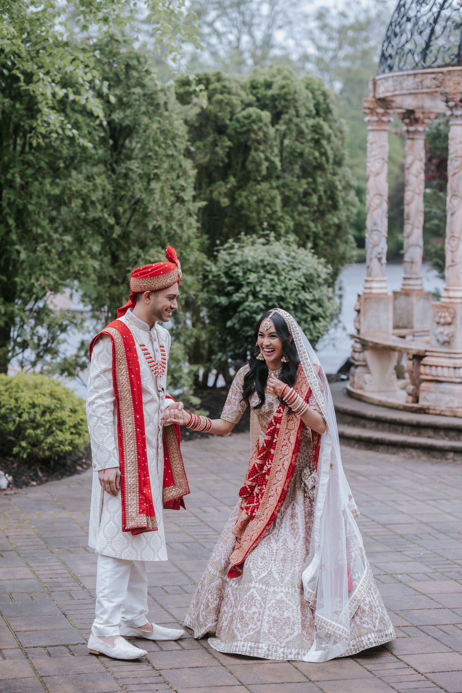 Indian bride and groom laughing together in traditional wedding outfits near stone gazebo.
