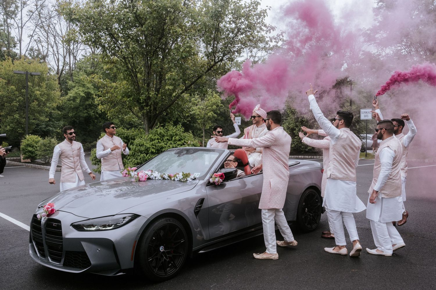 Groom arriving in a gray BMW convertible surrounded by groomsmen and pink smoke at baraat.