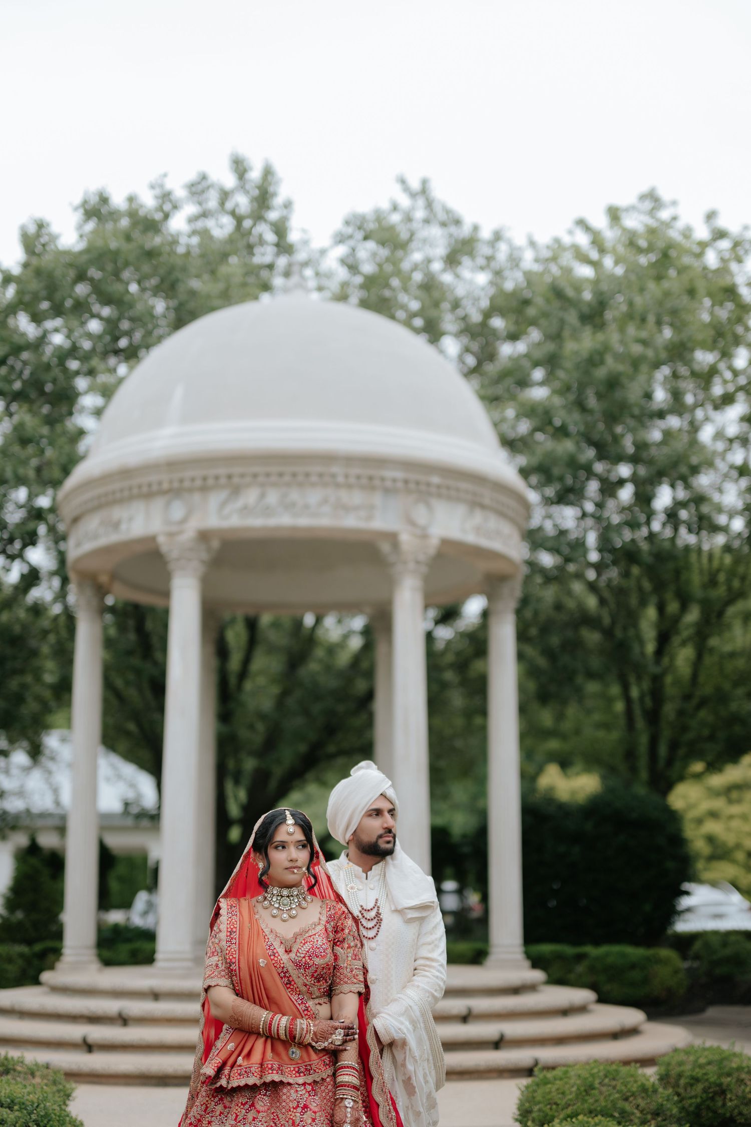 Indian bride and groom in traditional red and white wedding attire pose before an elegant white domed gazebo.