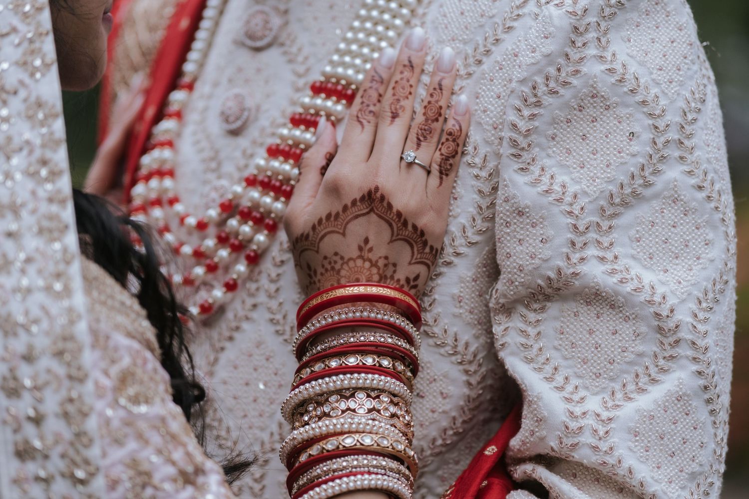 Close-up of Indian bride's hands adorned with intricate mehndi henna, red bangles, and pearl jewelry at wedding.
