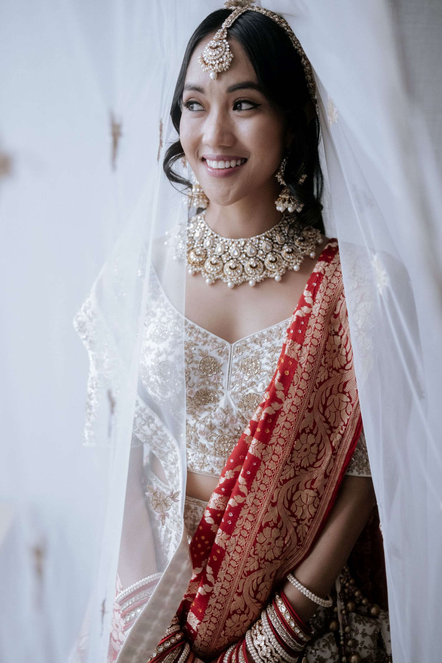 Smiling Indian bride in white and red lehenga with gold kundan jewelry and white veil on wedding day.