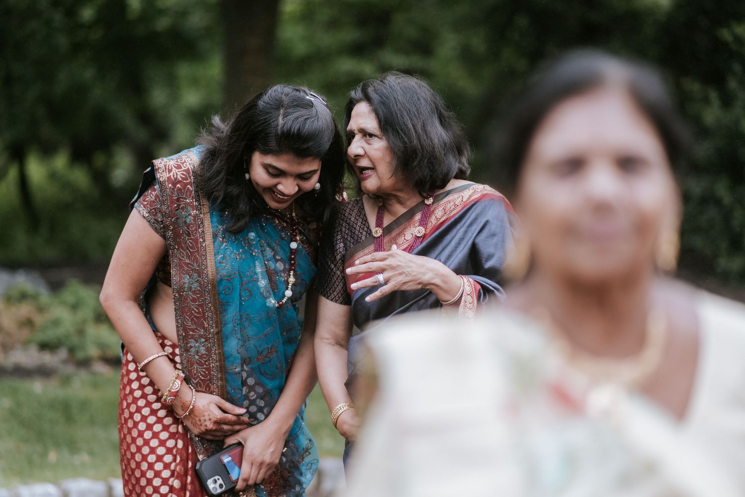 Two women in colorful Indian sarees sharing a warm moment together at an outdoor wedding.