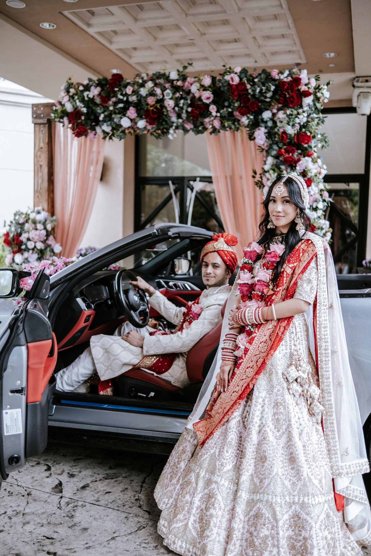 Indian bride in white lehenga standing beside groom in car under floral wedding arch decor.