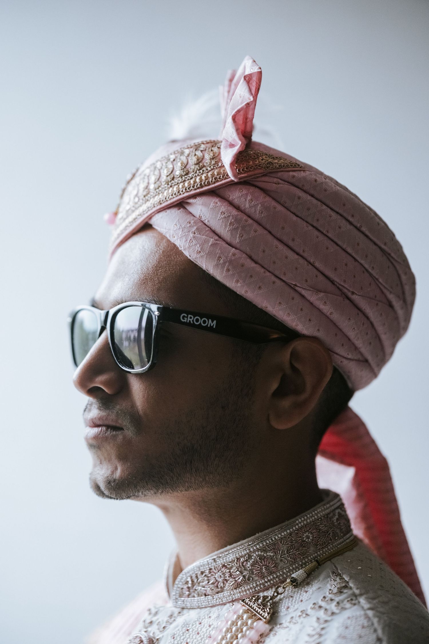 Indian groom wearing pink turban and sunglasses labeled GROOM poses in traditional sherwani for wedding portrait.