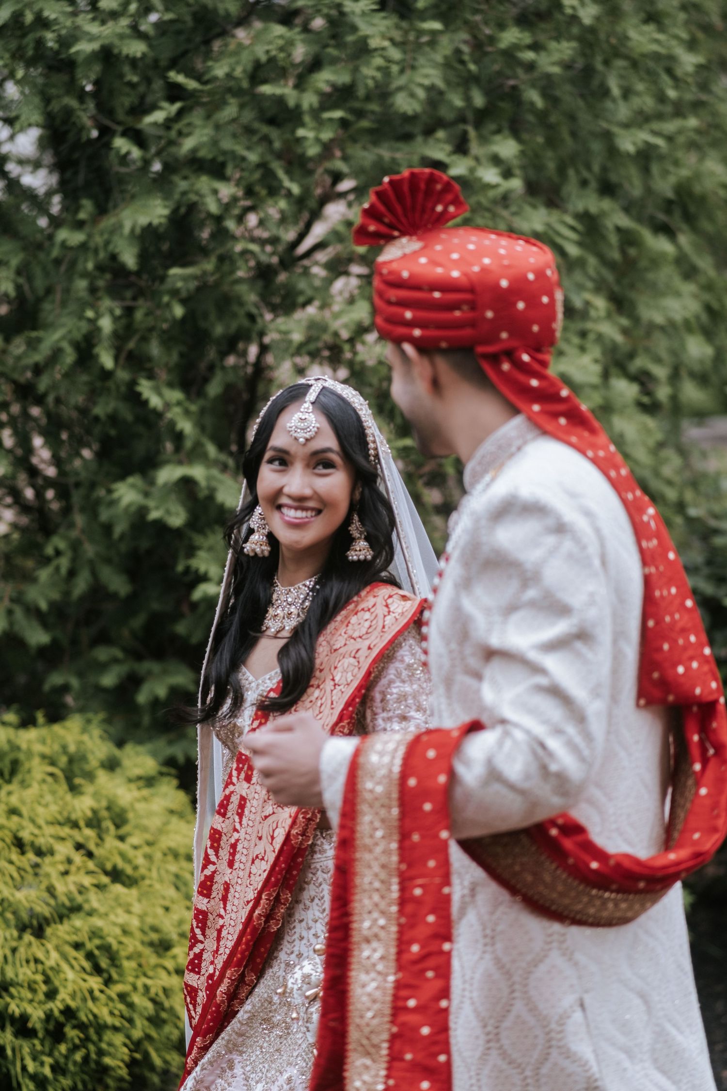 Smiling Indian bride in red and gold lehenga gazing at groom in white sherwani and red turban outdoors.