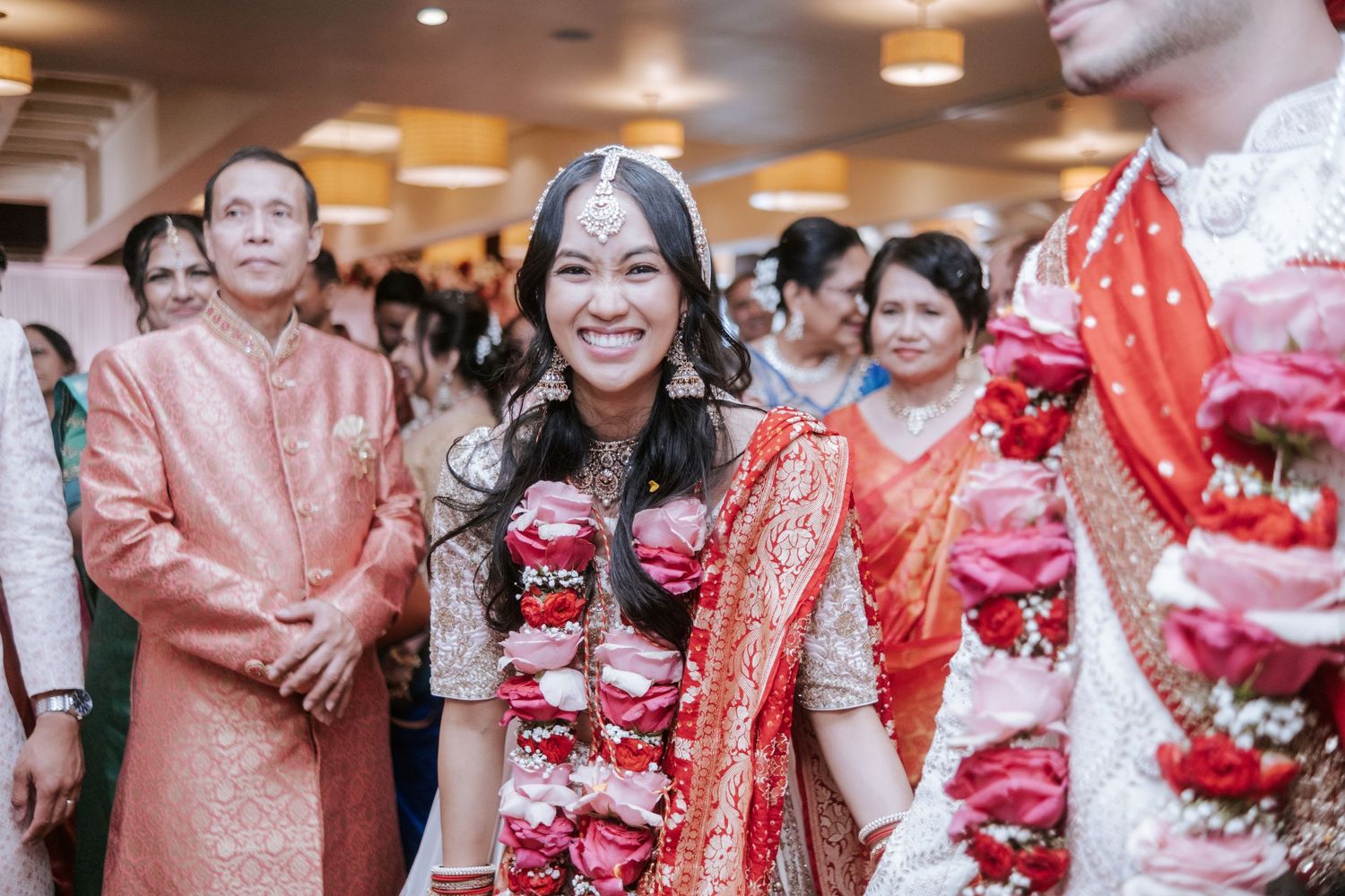 Radiant Indian bride in red and white lehenga smiles during traditional wedding ceremony procession.