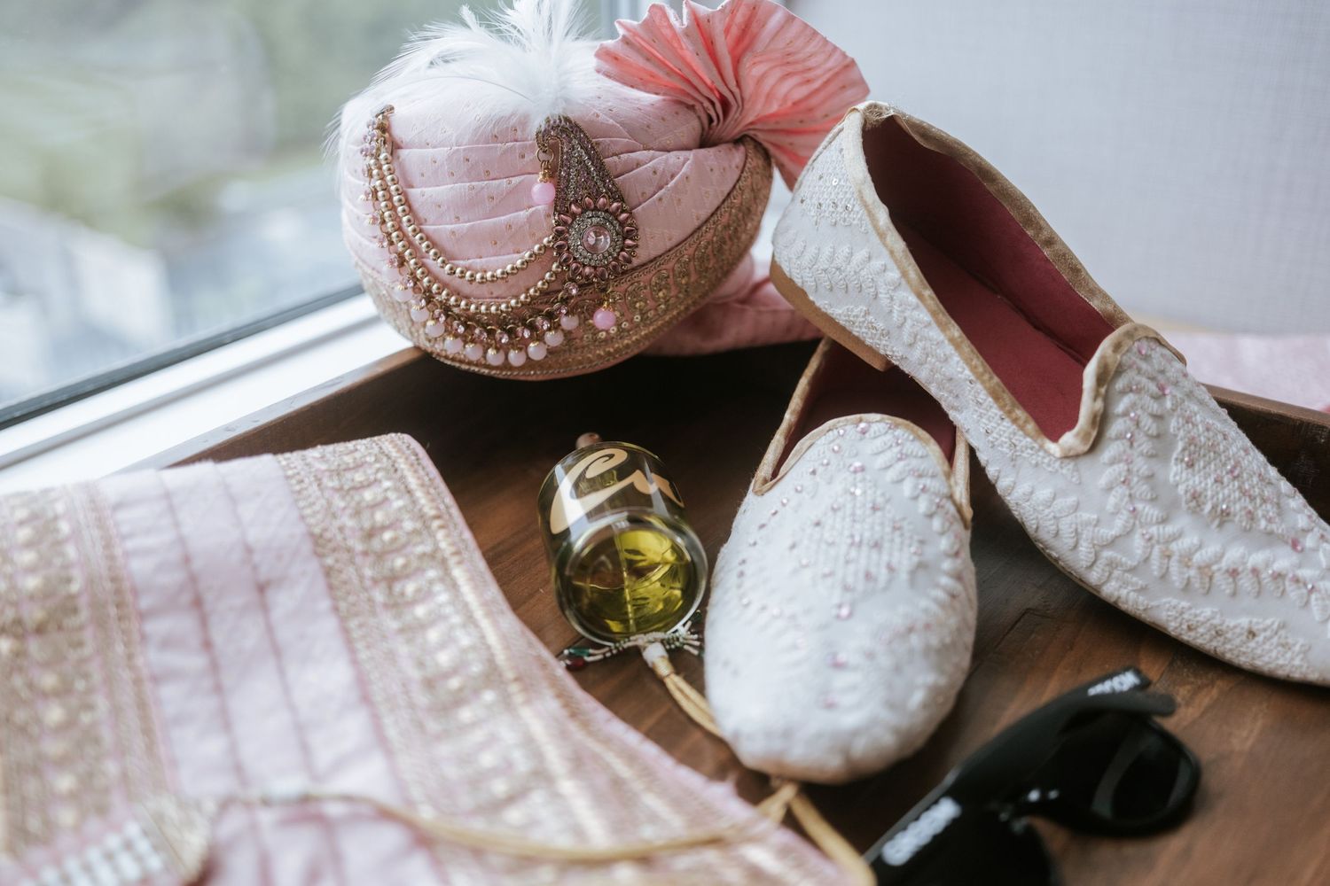 Indian wedding accessories including white embroidered juttis, pink turban, gold watch, and sunglasses on wooden surface.
