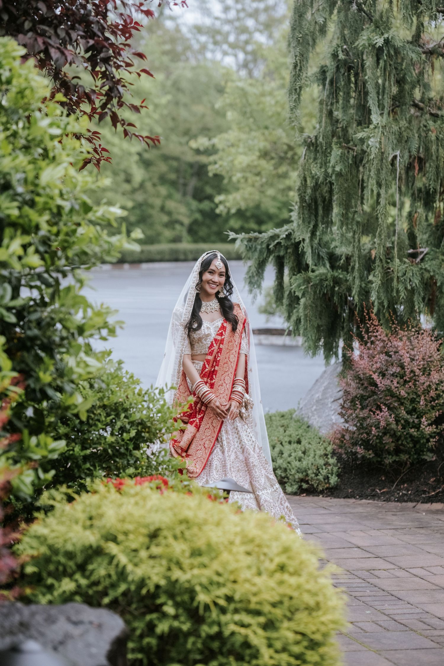 Elegant Indian bride in red and gold lehenga with white veil poses gracefully in a lush garden setting.