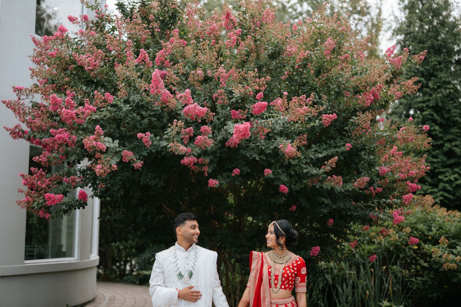 Indian wedding couple in traditional red and white attire posing near blooming pink trees.