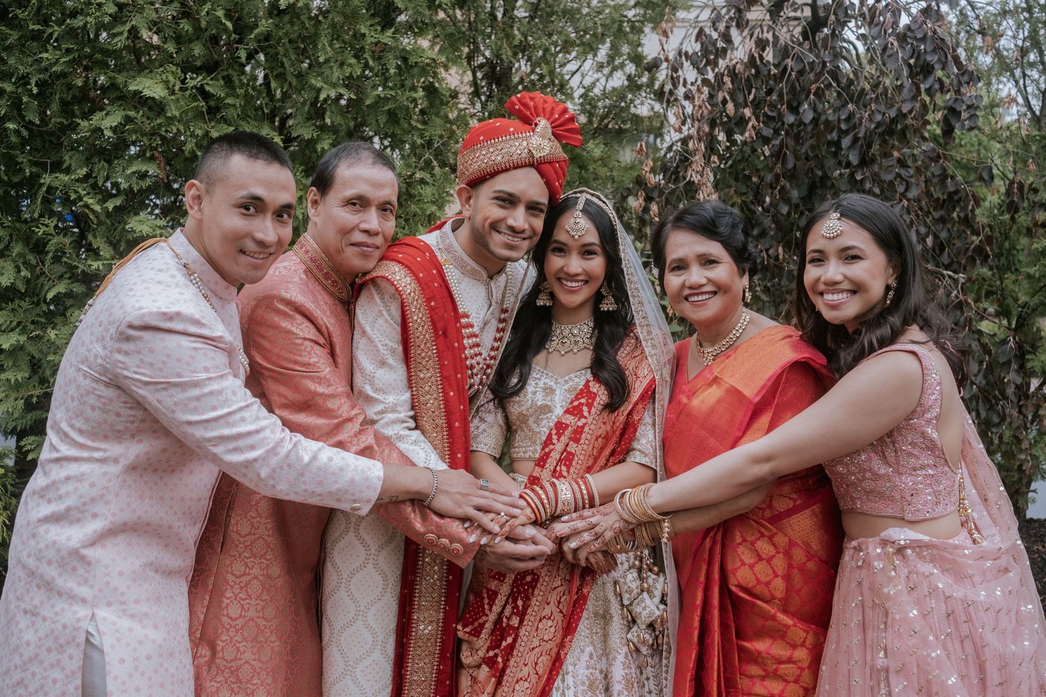 Indian bride and groom celebrate with wedding party in traditional South Asian attire, joining hands together.
