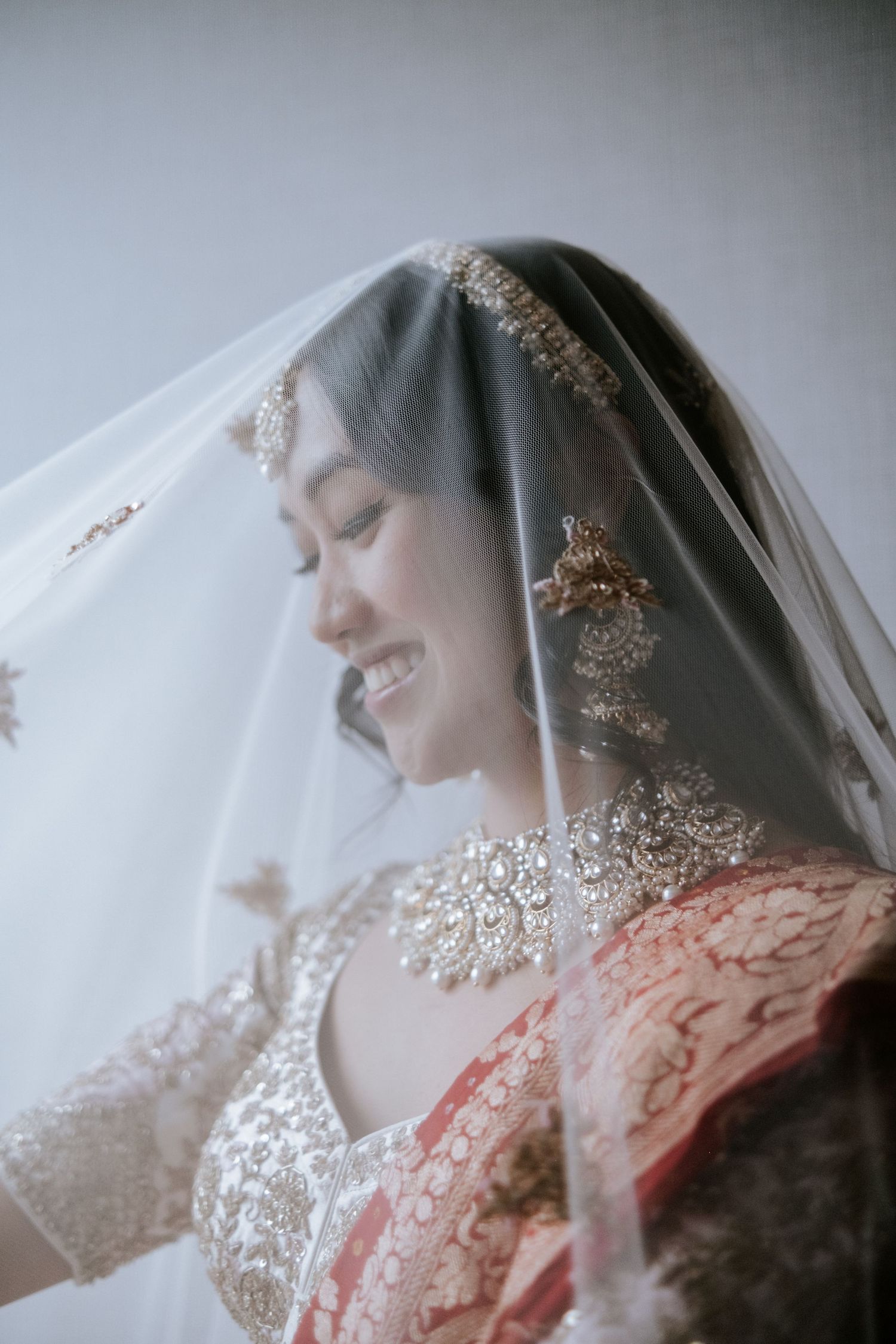 Smiling Indian bride covered by delicate veil wearing gold chandelier earrings and lehenga.