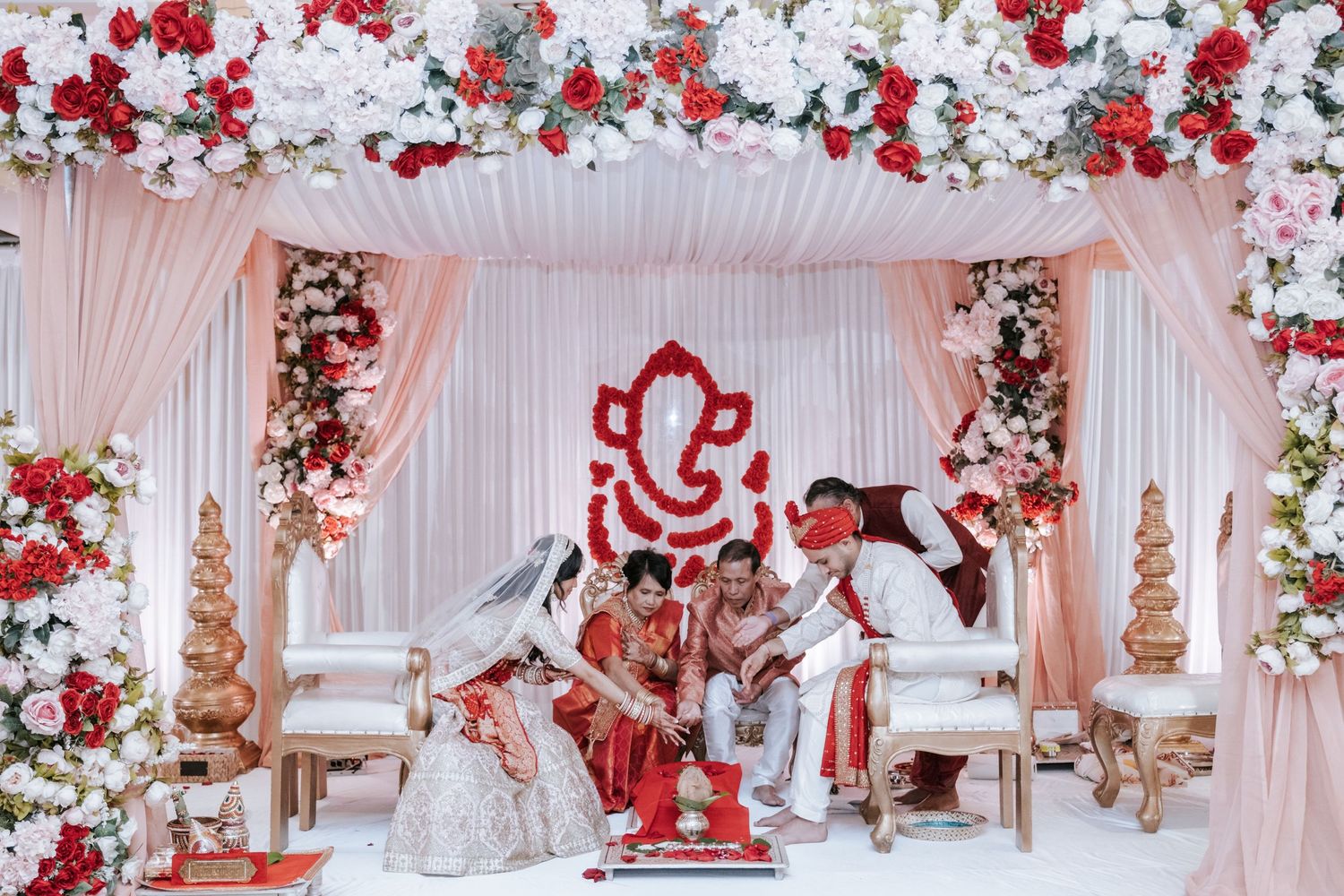 Bride and groom seated at ornate Hindu wedding mandap with red Ganesh and floral decor.