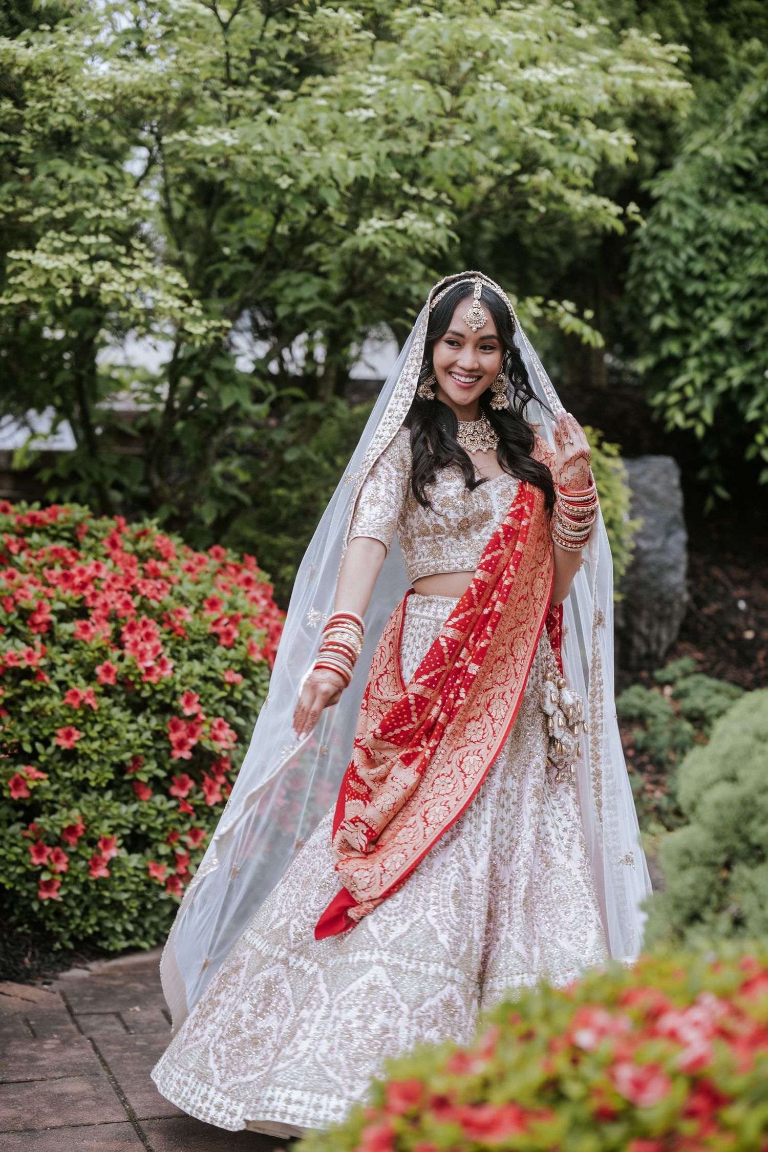 Indian bride in white and red bridal lehenga twirling in garden with floral backdrop.