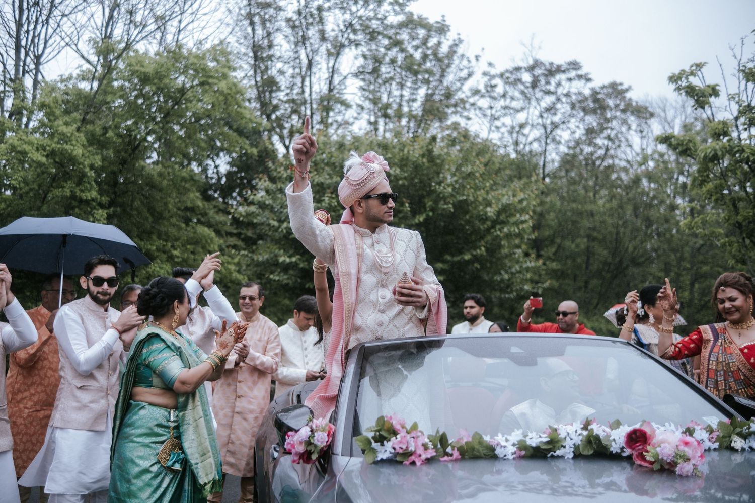 Indian groom in pink sherwani celebrating joyfully atop a decorated car during the baraat procession.