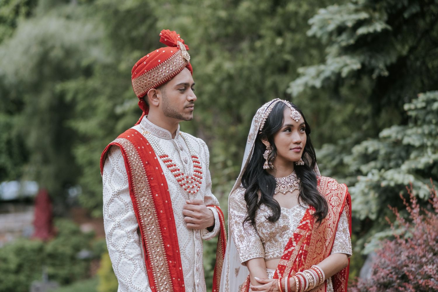 Indian wedding couple in traditional attire, groom in white sherwani and red turban with bride in lehenga outdoors.