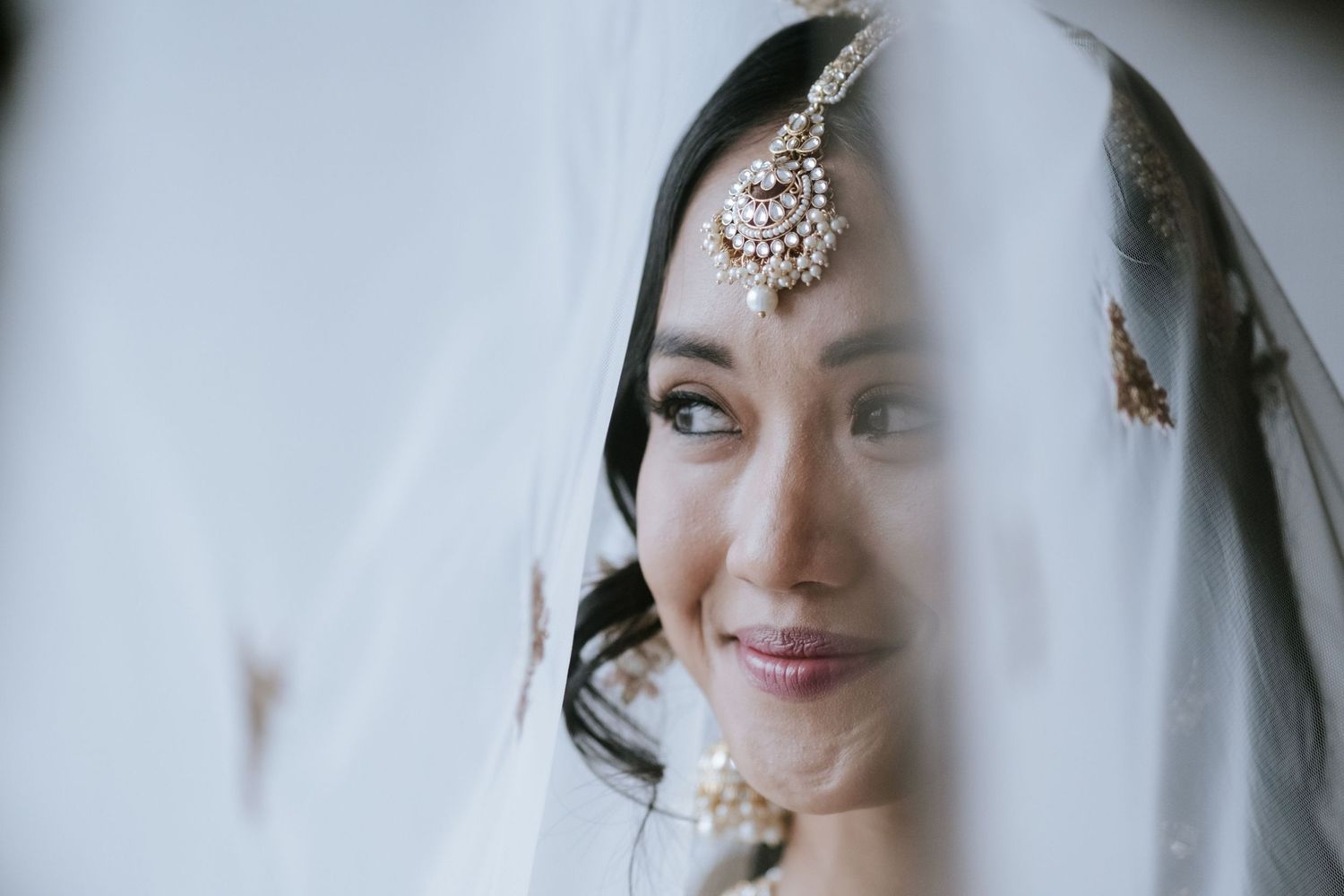 Close-up of Indian bride peeking through white veil wearing maang tikka bridal jewelry.