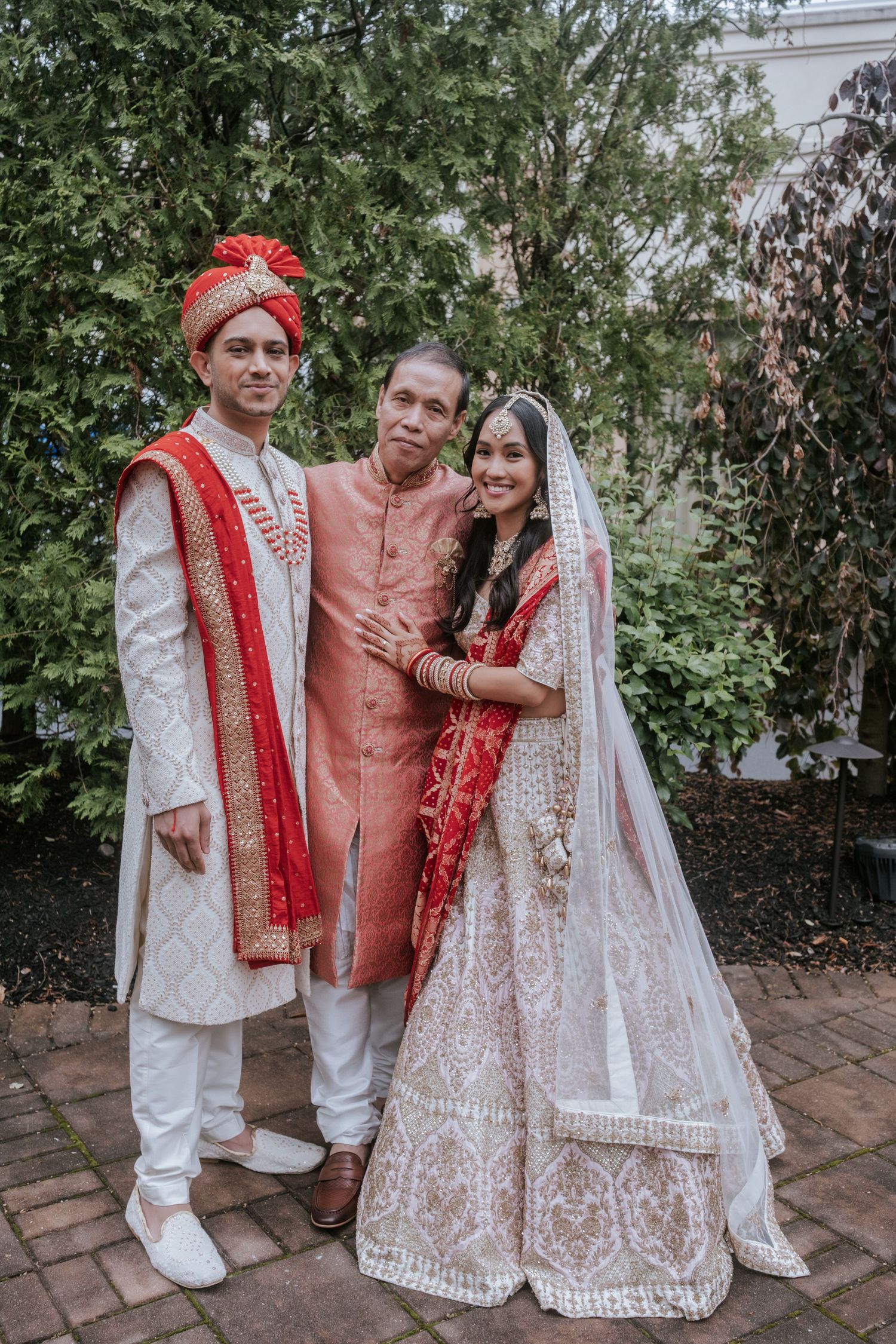 Indian groom in white sherwani and bride in red lehenga pose with older man in garden setting.