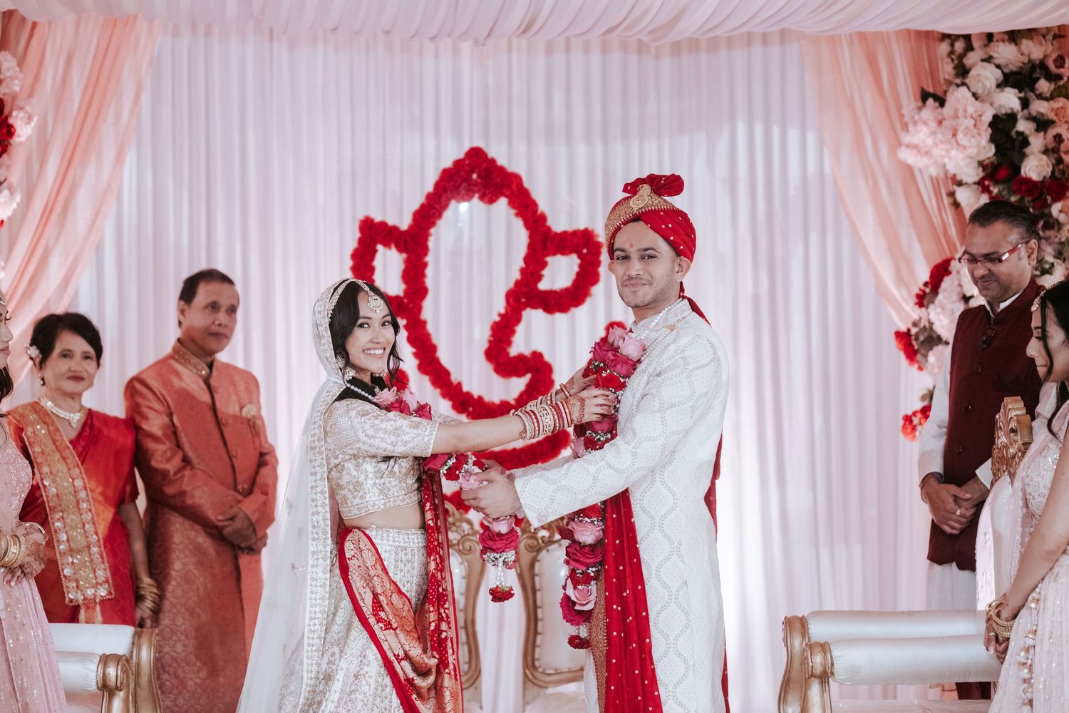 Bride and groom exchanging garlands at Hindu wedding ceremony with pink floral mandap backdrop.