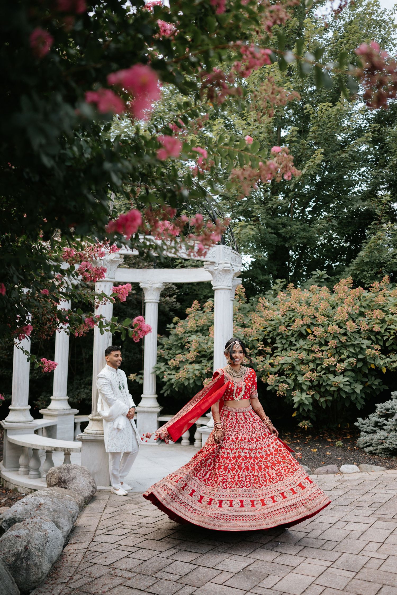 Indian bride in red lehenga twirling near white gazebo with groom in white sherwani at outdoor wedding.