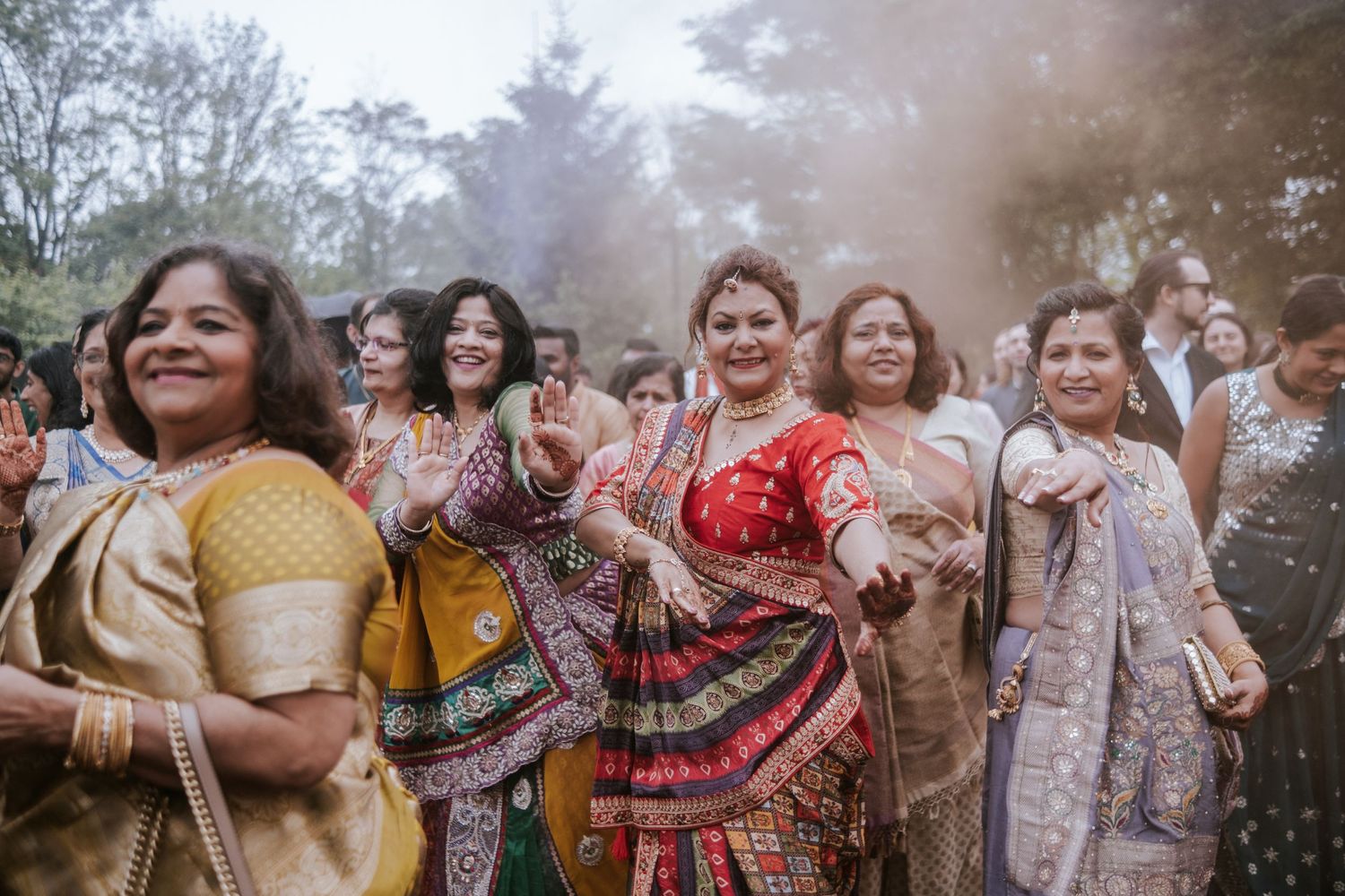Women in vibrant Indian sarees dancing joyfully during a colorful outdoor wedding celebration.