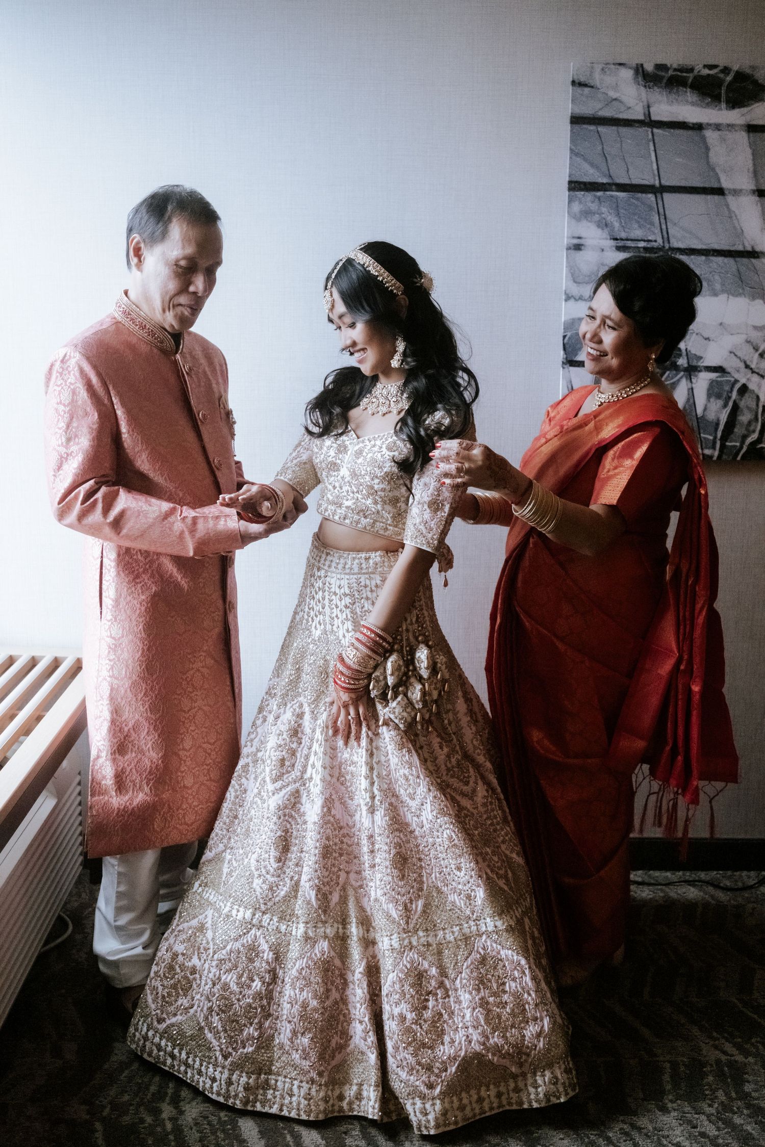 Indian bride in ivory lehenga shares a tender moment with her parents during wedding day preparations.