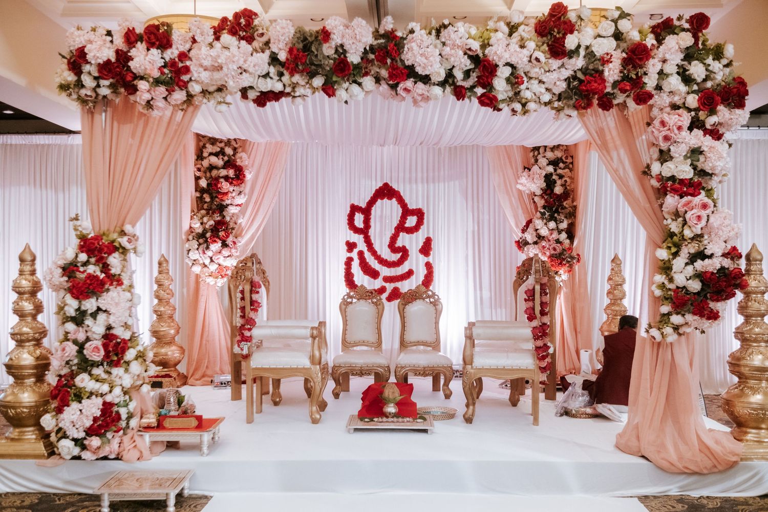Elaborate Indian wedding mandap decorated with red and white flowers, gold pillars, and Ganesha centerpiece.
