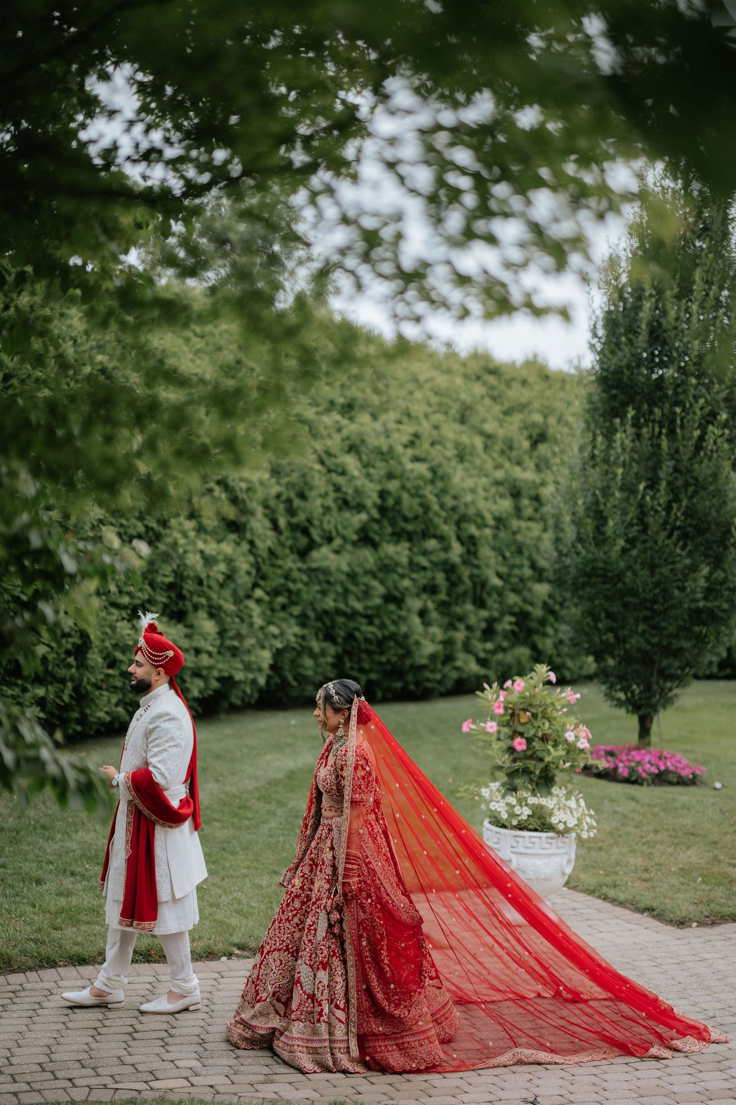 Indian bride in stunning red lehenga with flowing veil walks with groom through lush garden path.