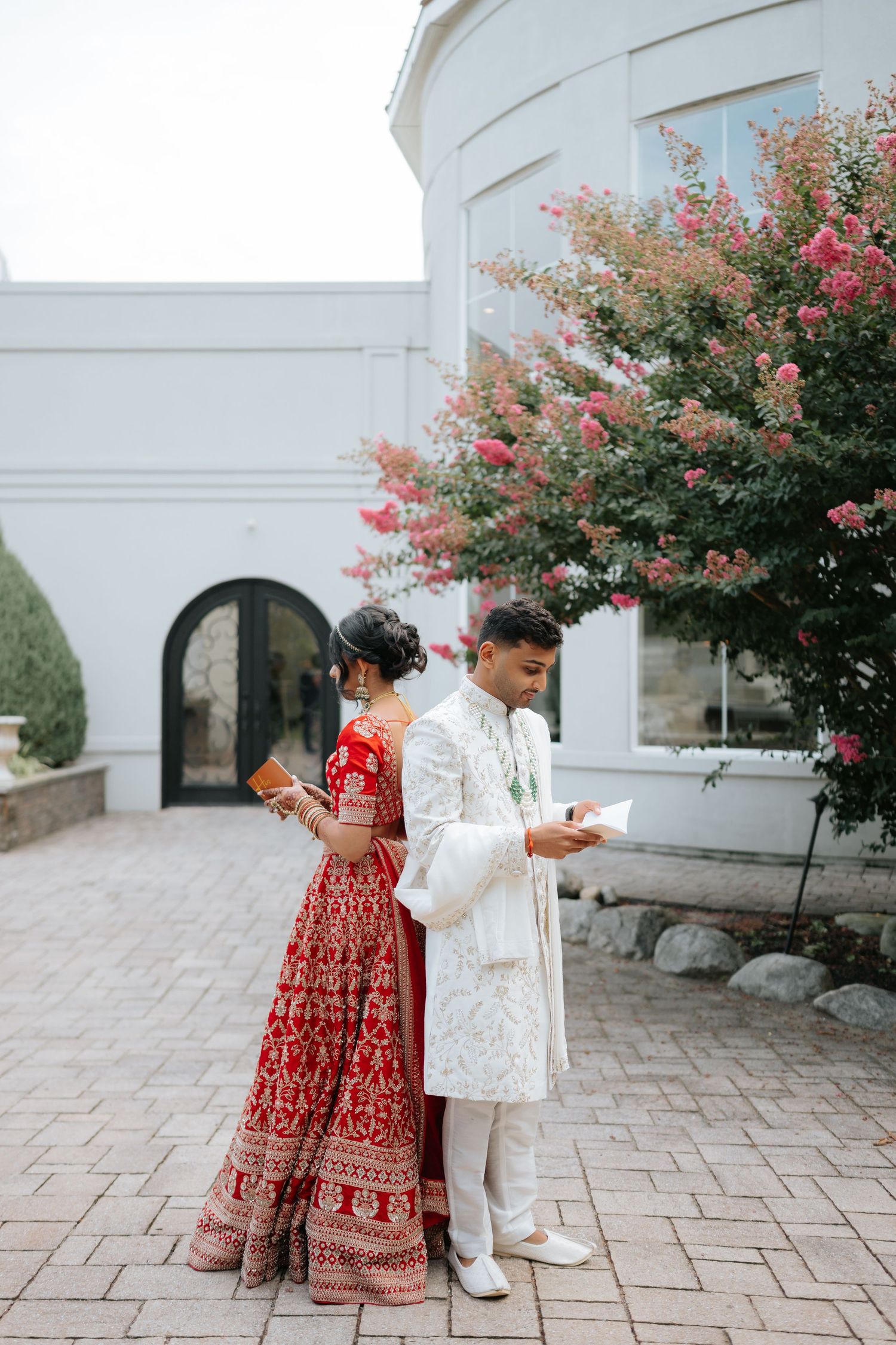 Indian bride and groom standing back-to-back reading vows outside elegant white wedding venue.
