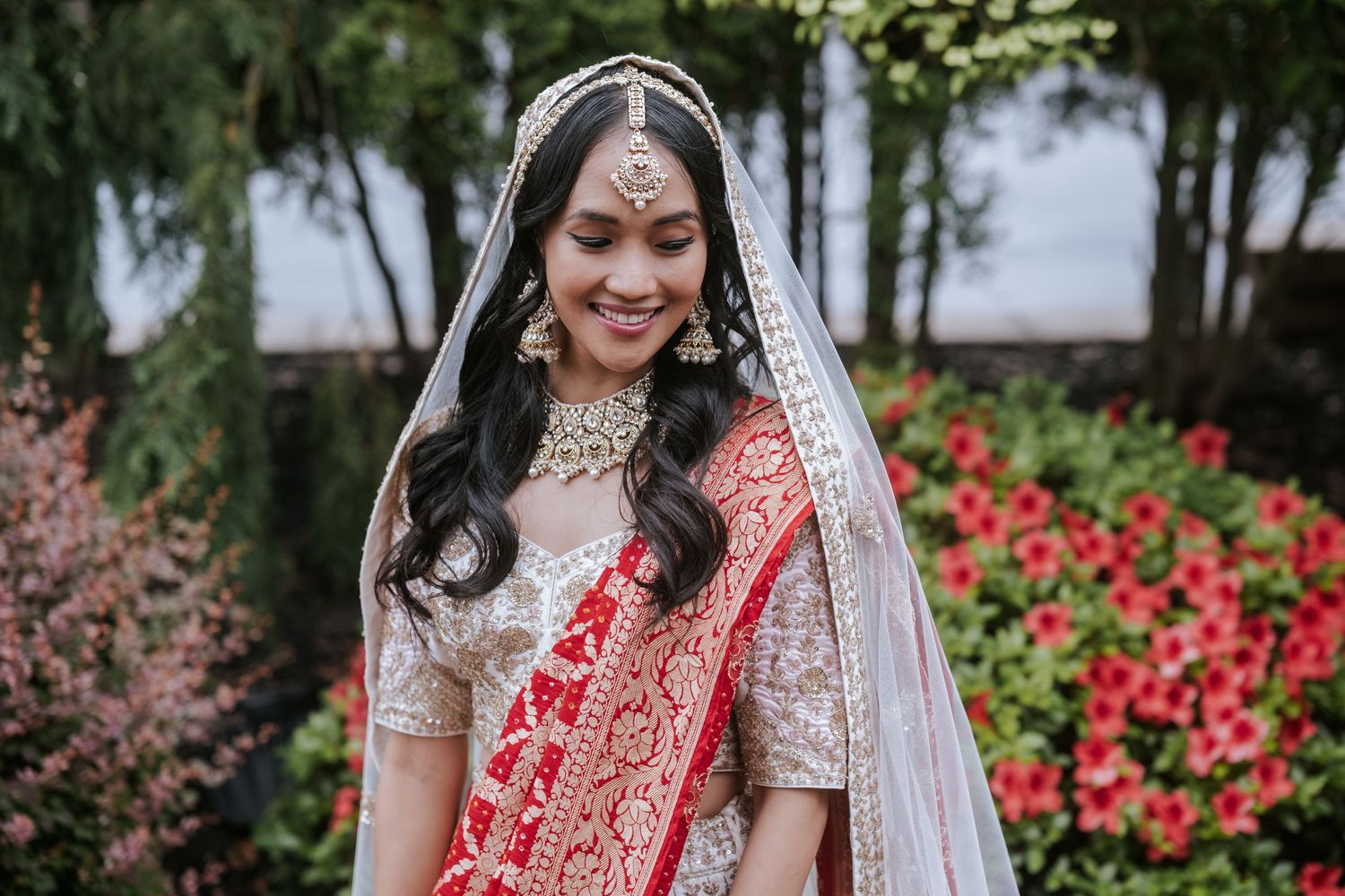 Indian bride in white and red lehenga with gold jewelry and veil smiling outdoors.