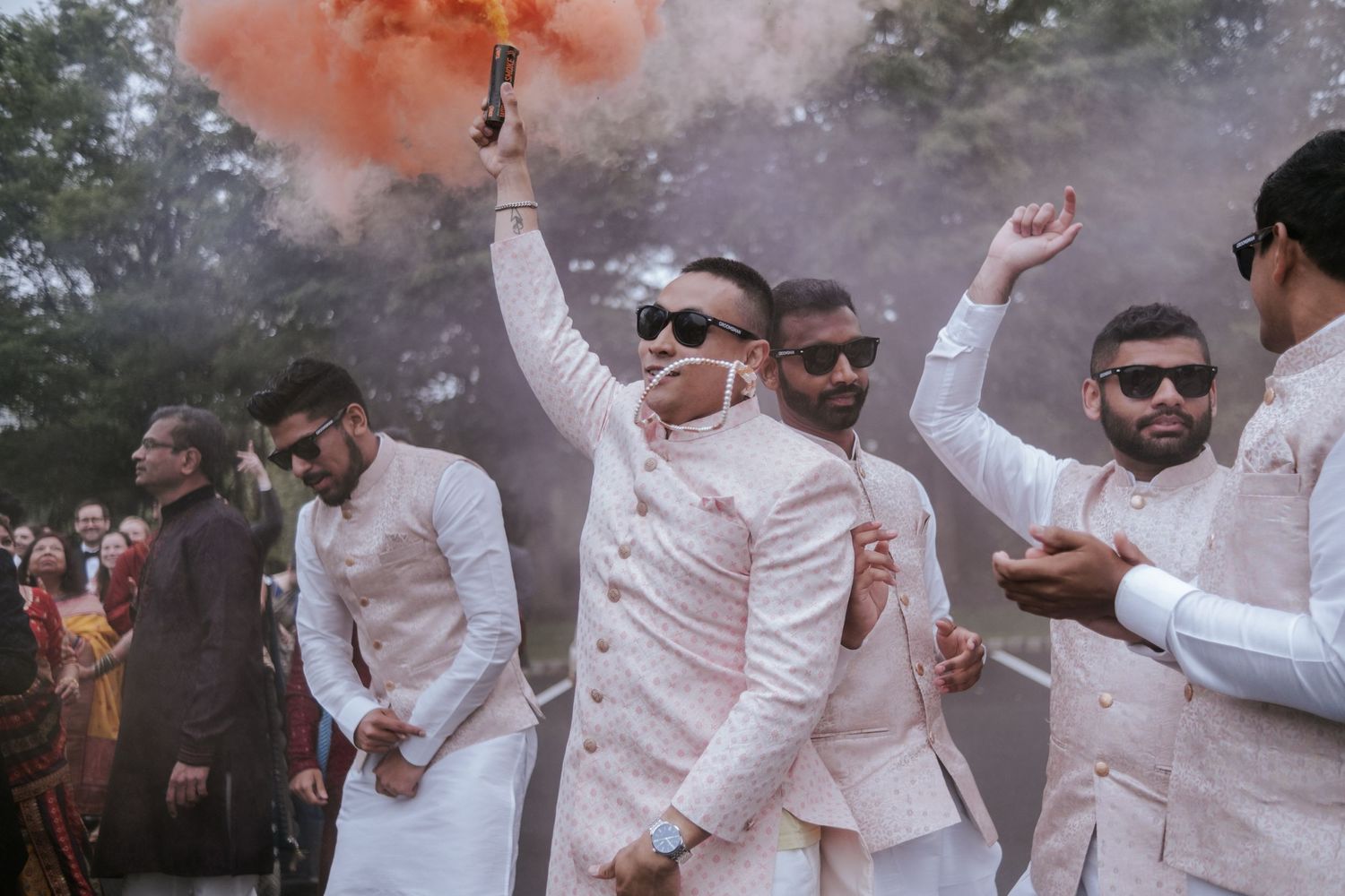 Groom in white sherwani holding orange smoke flare during Indian wedding baraat procession.