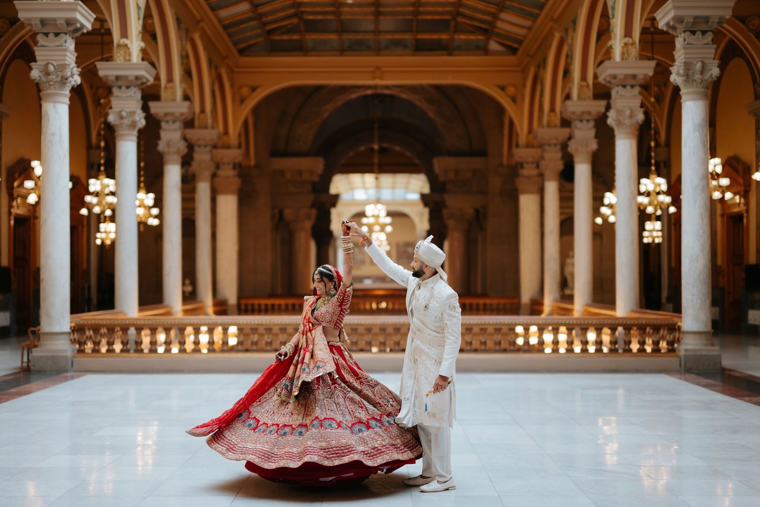 Indian bride in red lehenga twirling with groom in white sherwani in ornate golden hall.