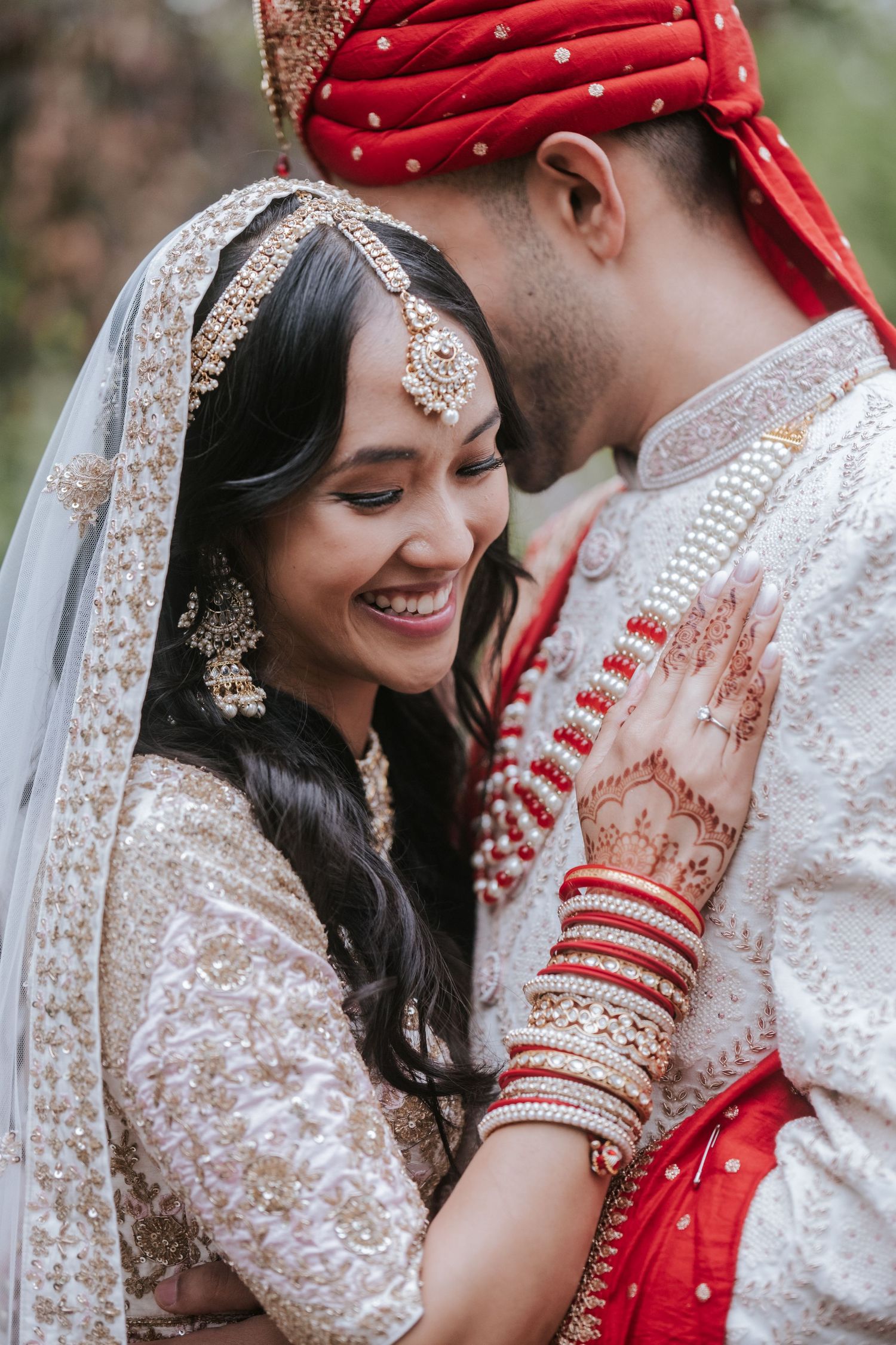 Smiling Indian bride in white lehenga with mehndi hands resting on groom's chest during outdoor wedding portraits.