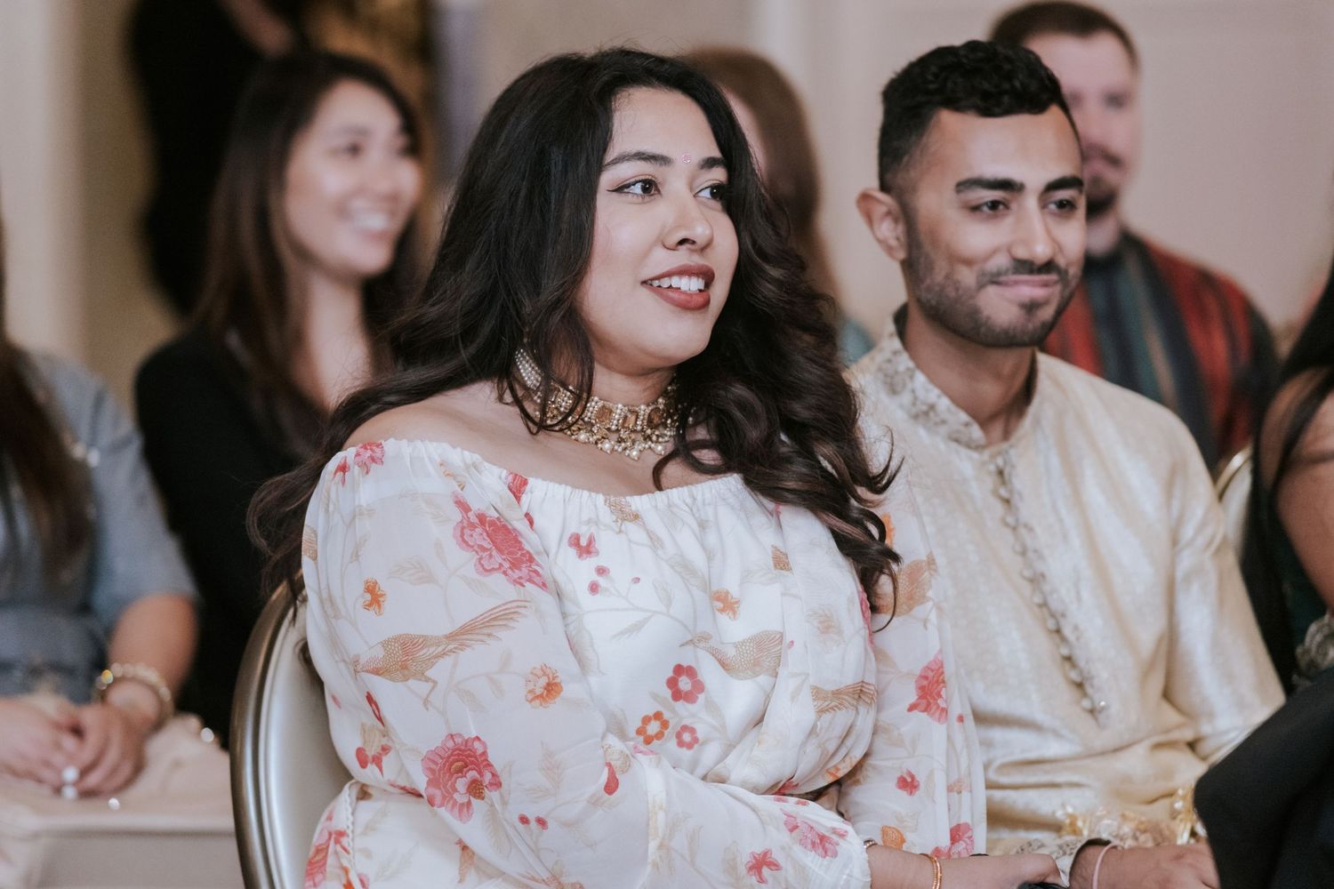 Woman in floral off-shoulder dress smiles while seated at an Indian wedding ceremony indoors.