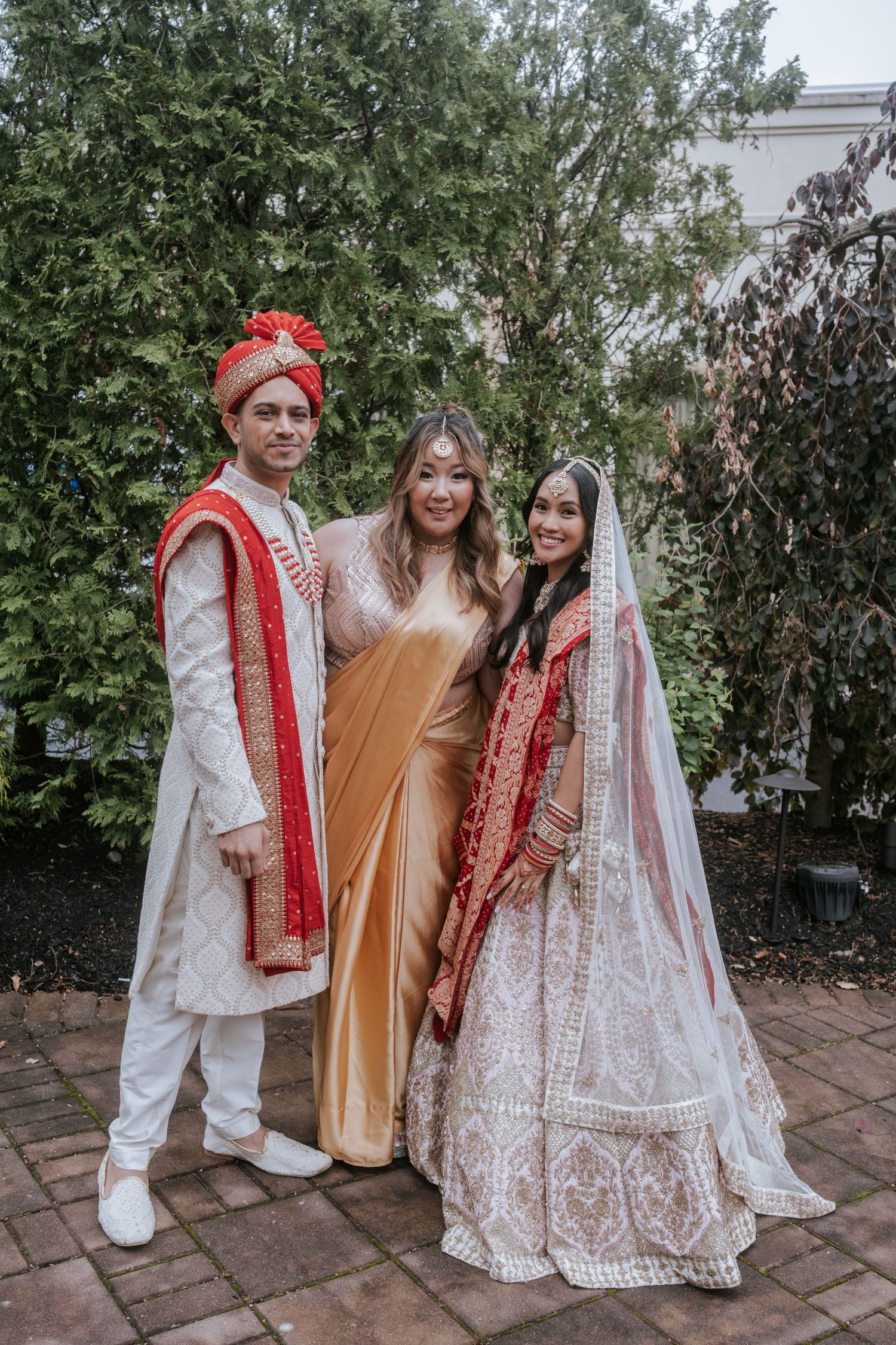 Indian groom in white sherwani and bride in red lehenga pose with woman in golden saree outdoors.