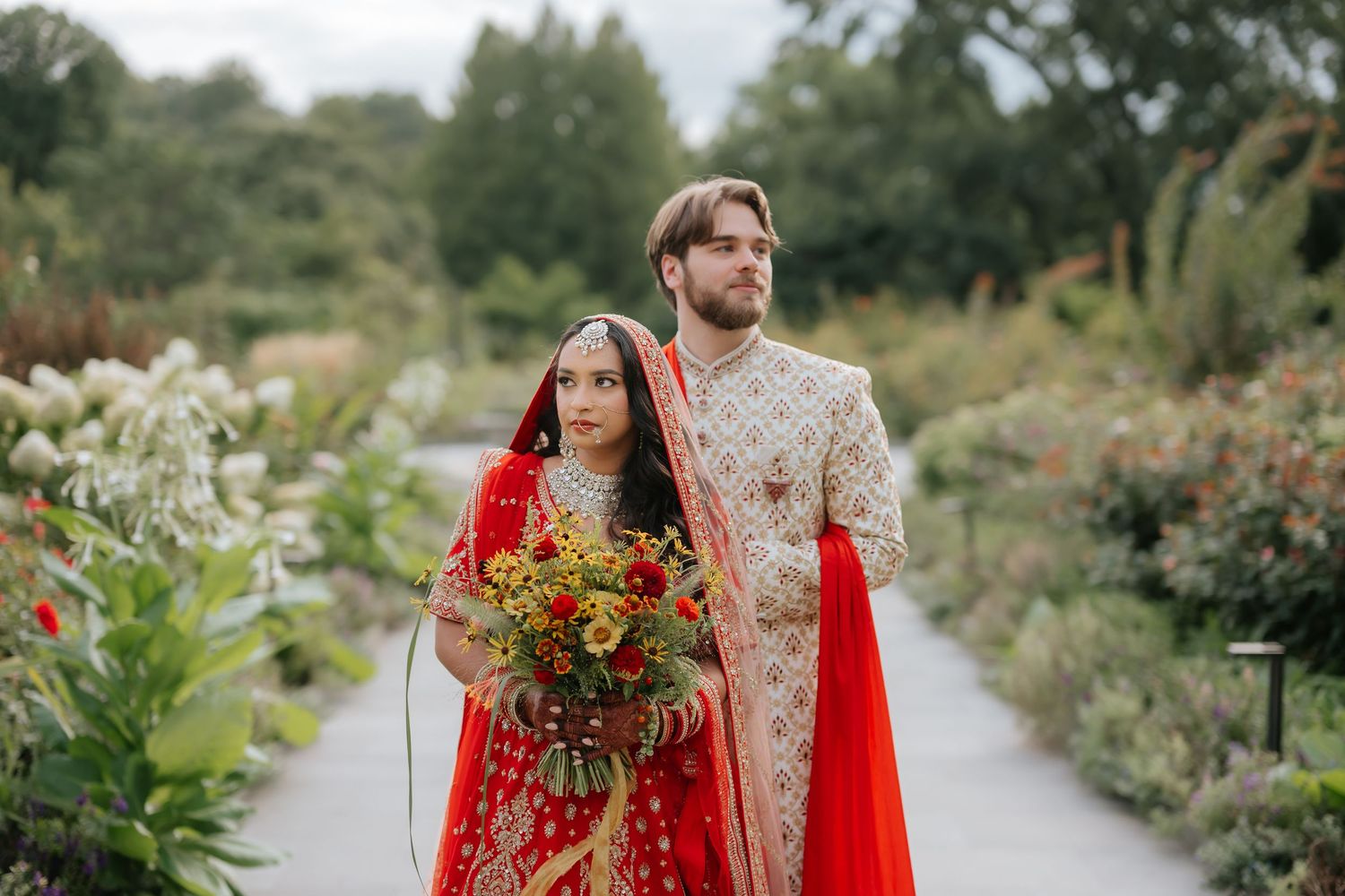 Indian bride in red lehenga holding colorful bouquet with groom in sherwani in garden setting.