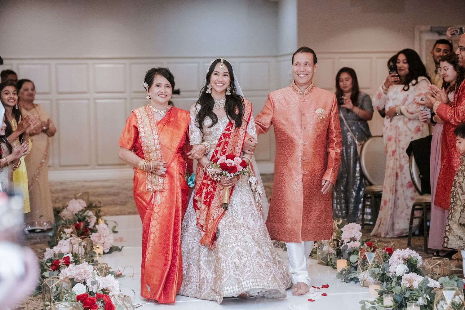 Bride in bridal lehenga walks down aisle with family at elegant Indian wedding ceremony.