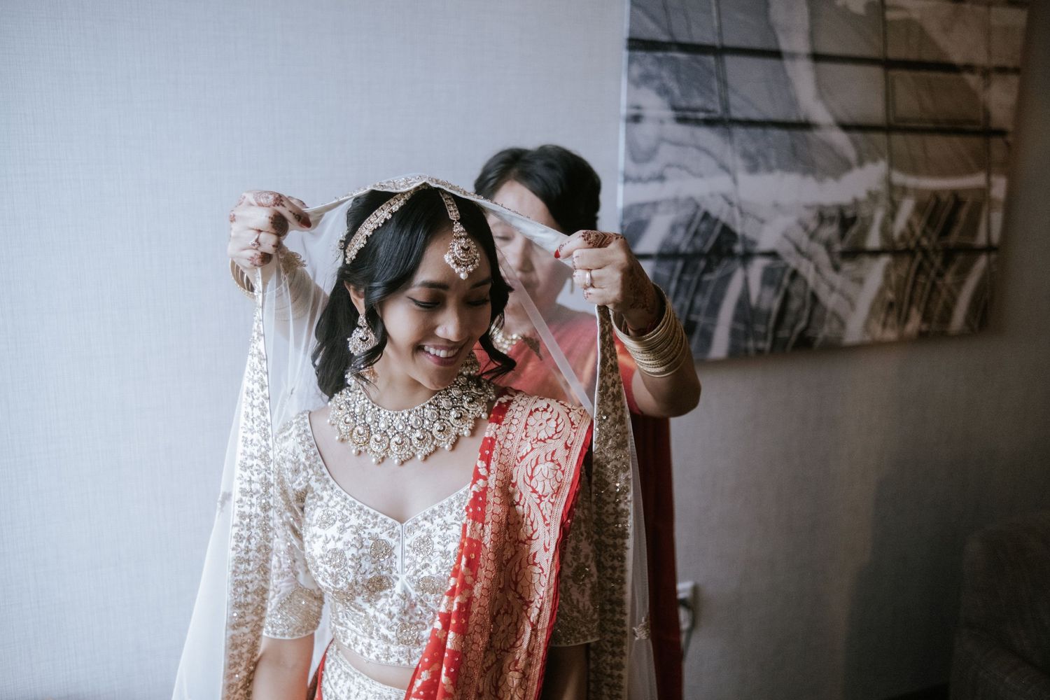 Indian bride smiles as veil is placed over her head during bridal preparations.