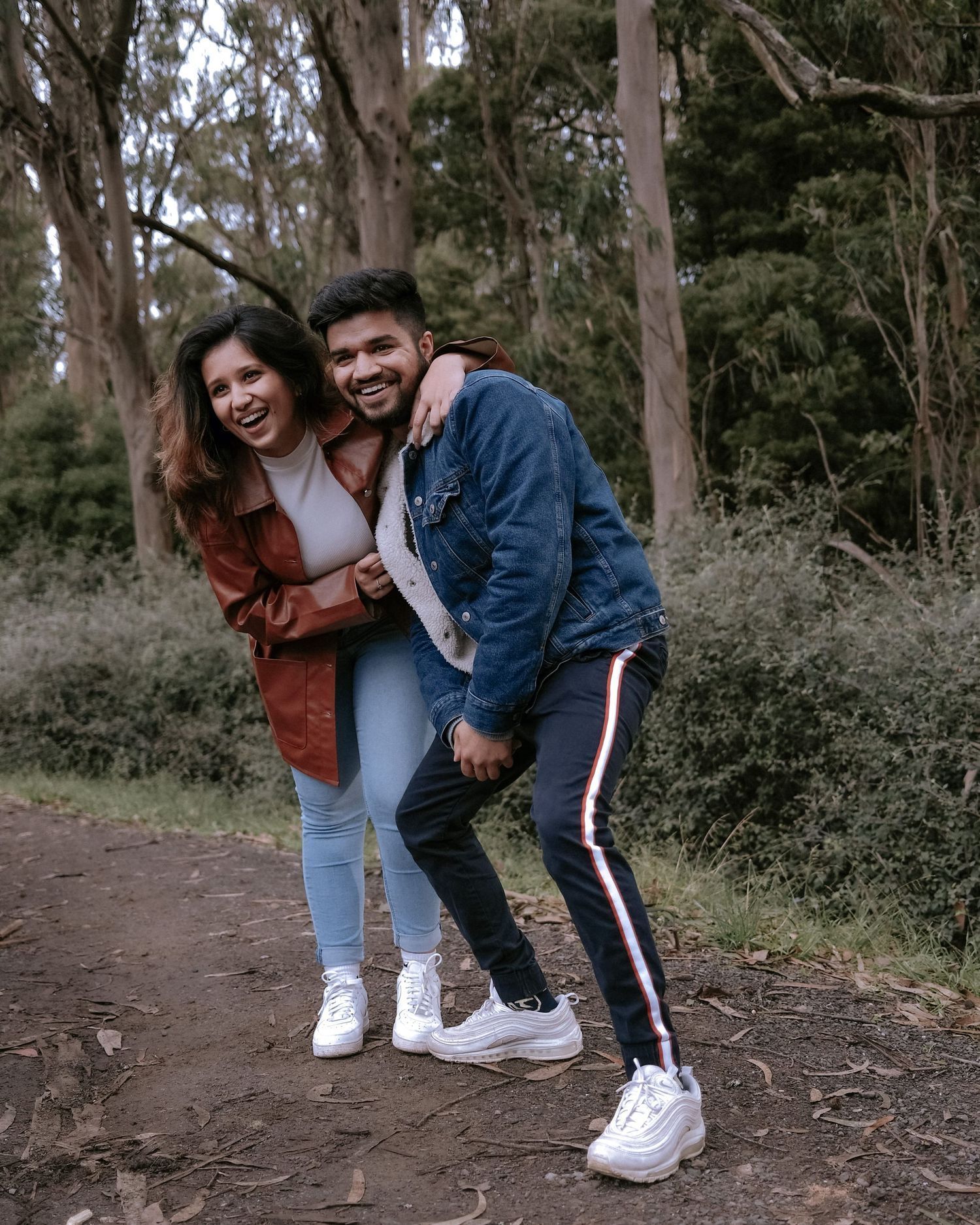 Happy South Asian couple laughing and embracing on a forest path during casual outdoor photoshoot.