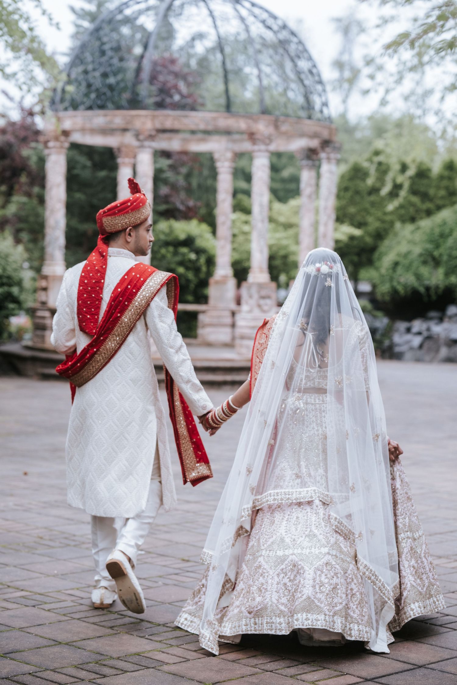 Indian wedding couple walking hand in hand toward stone gazebo, bride in white lehenga with veil, groom in sherwani.